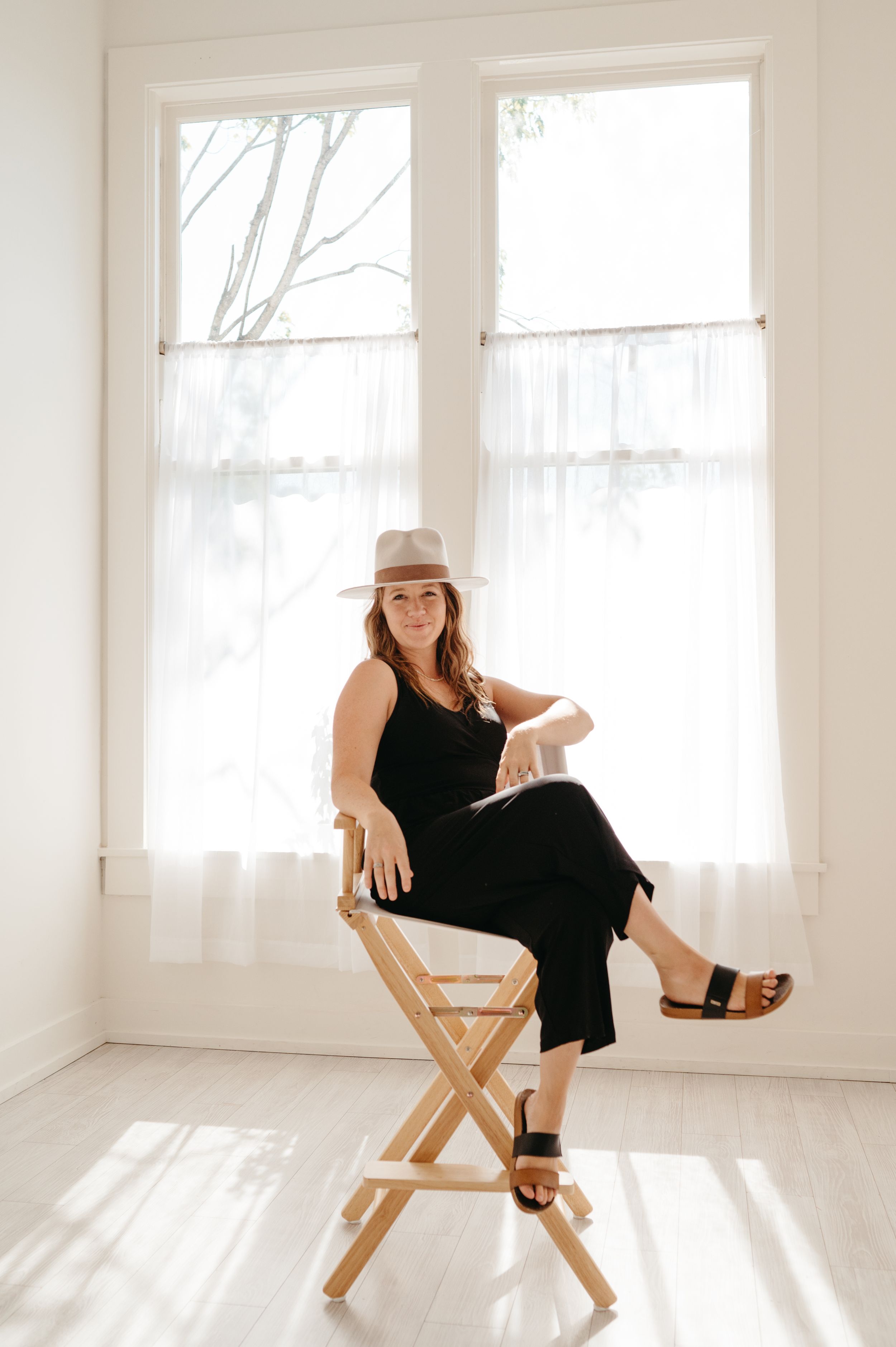 A person wearing a black dress and tan hat sits on a wooden director's chair in a bright white room with sheer curtains.