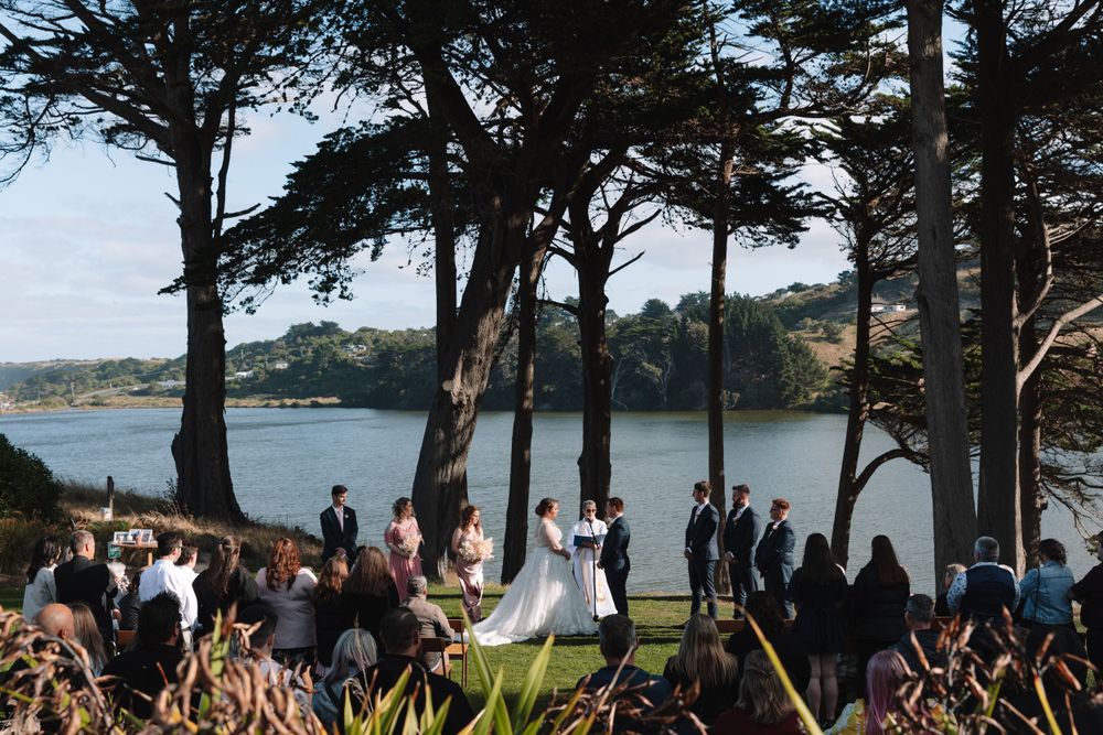 Wedding ceremony guests gather under tall trees overlooking a scenic bay during an outdoor celebration.