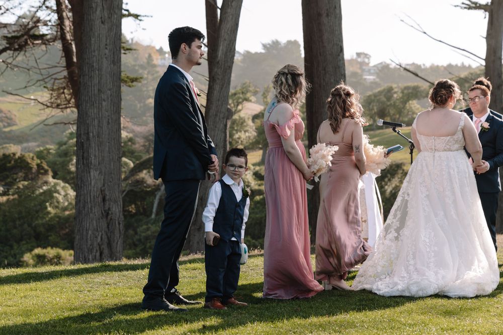 Wedding party in formal attire stands on a grassy hill with trees and coastal views.