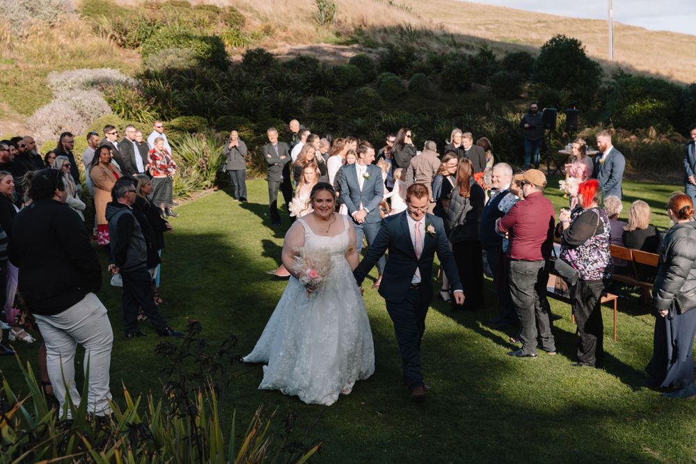 Wedding party walks down a grassy hillside path as guests look on during an outdoor celebration.
