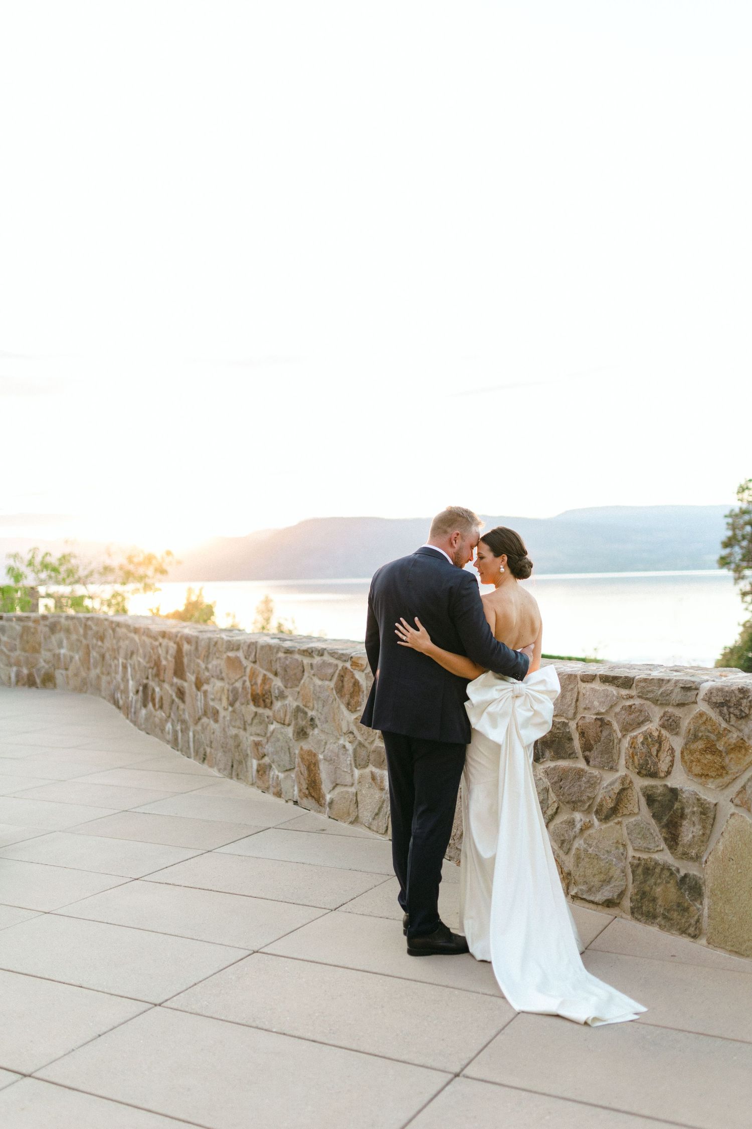 Couple embraces while overlooking scenic mountain landscape from stone wall terrace during sunset wedding moment.