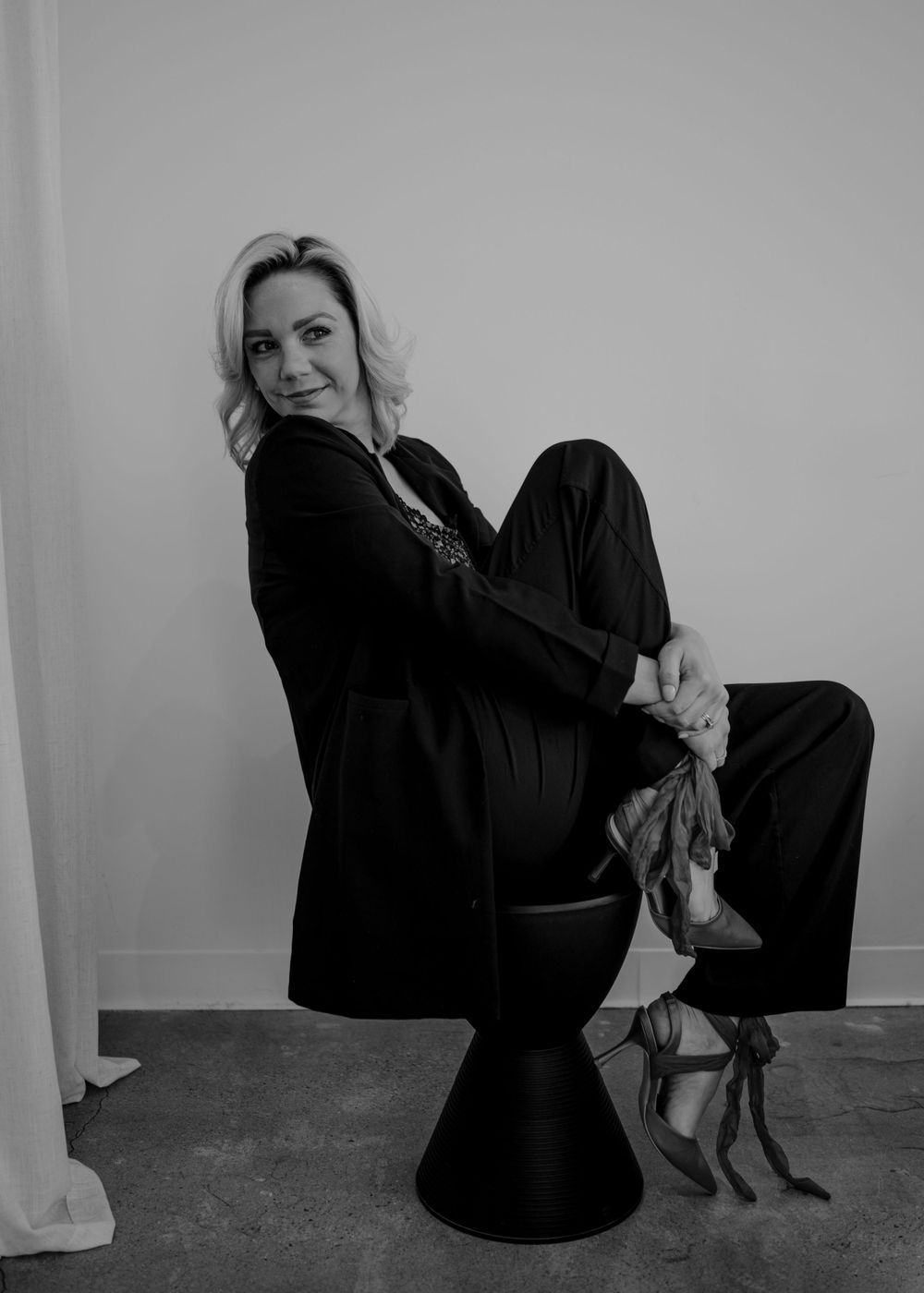 Stylish portrait of person in black outfit sitting on modern stool against light wall.