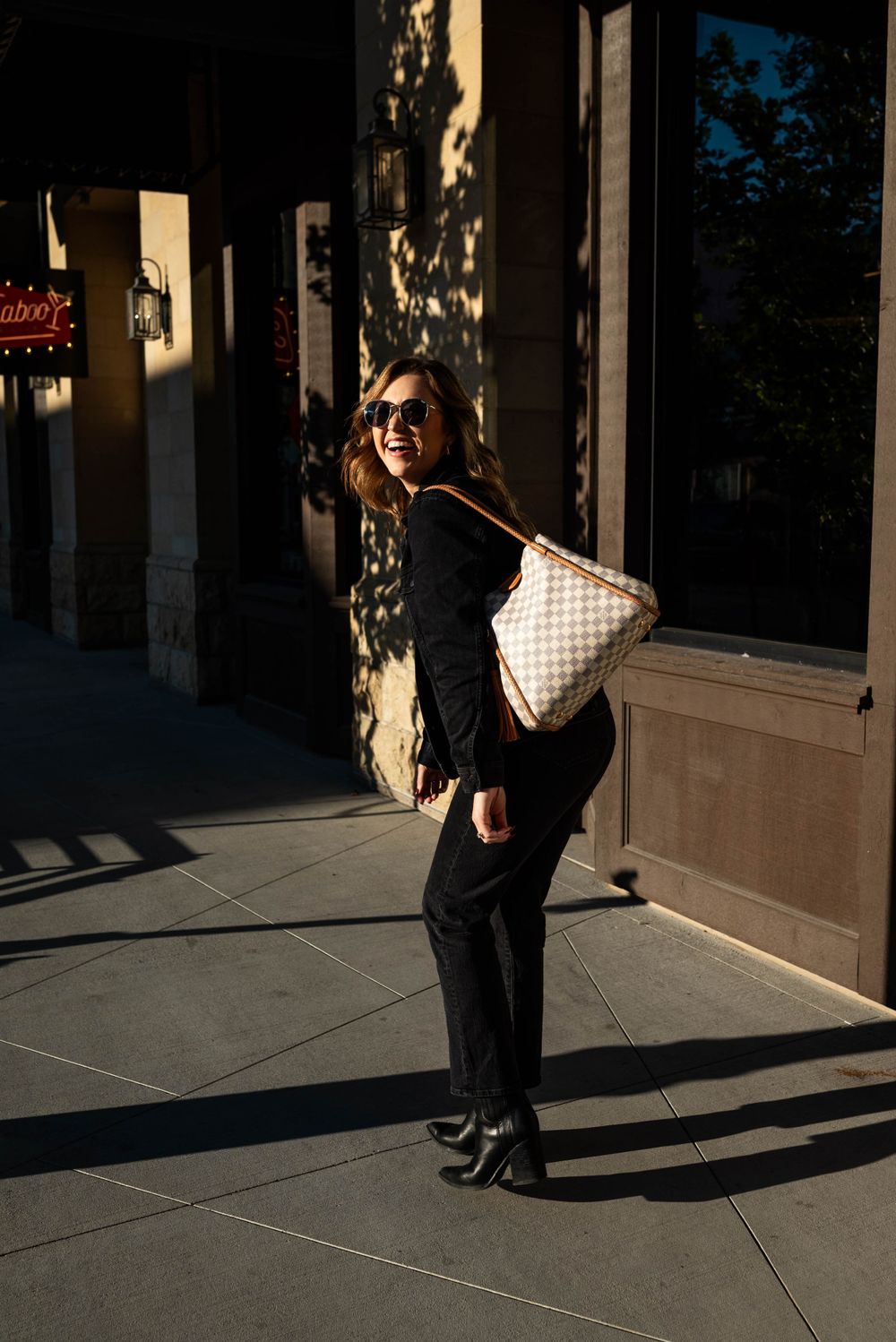 Stylish woman with checkered handbag poses on urban sidewalk in afternoon shadows.