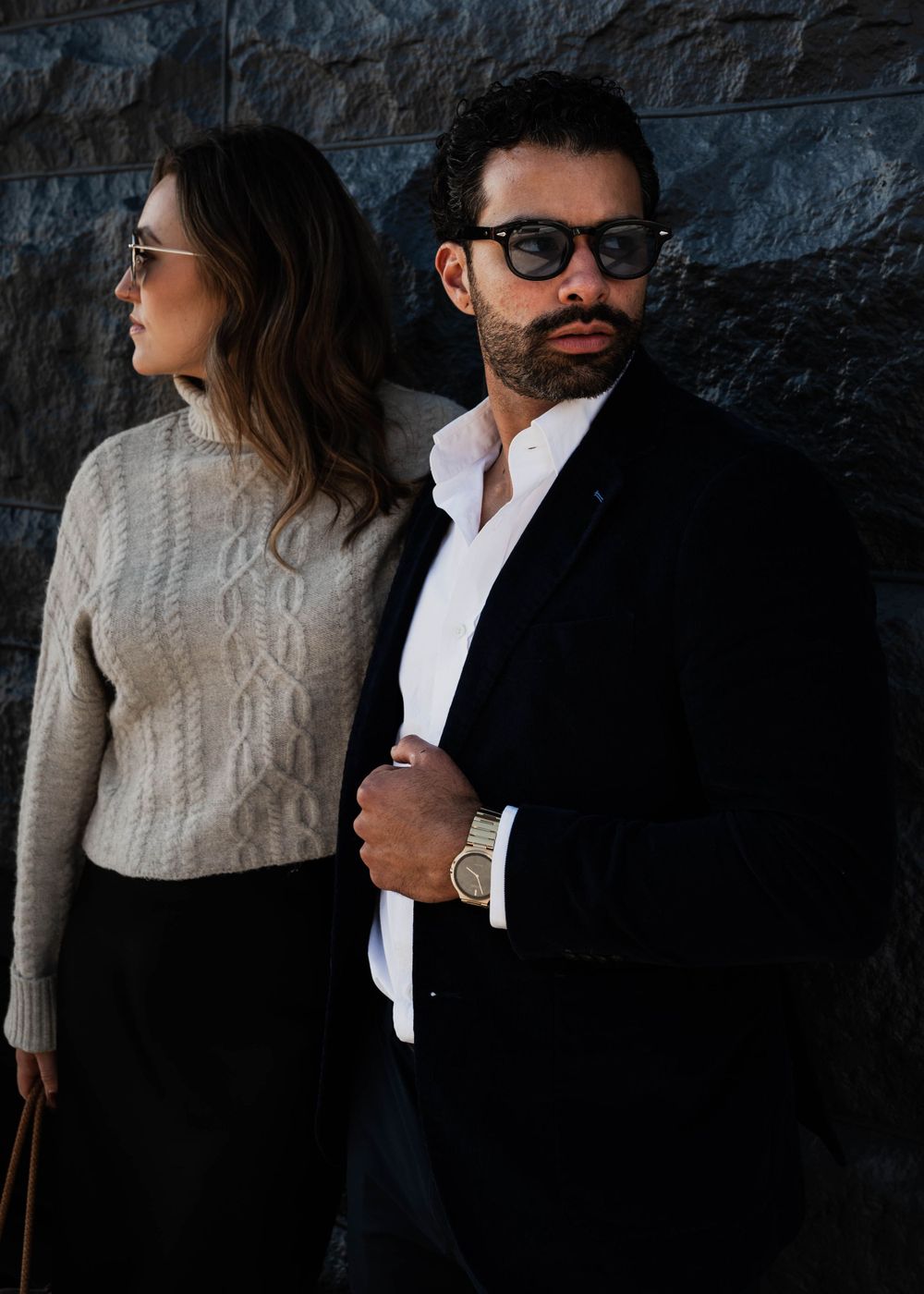 Fashionable couple in winter attire pose against textured stone wall background.