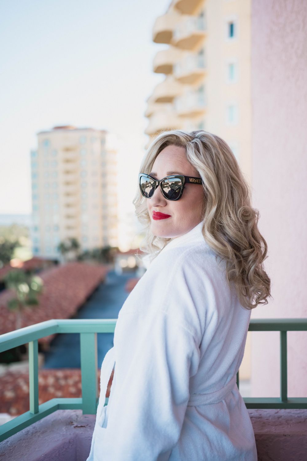 Glamorous woman in white coat and sunglasses poses on balcony with city buildings in background.
