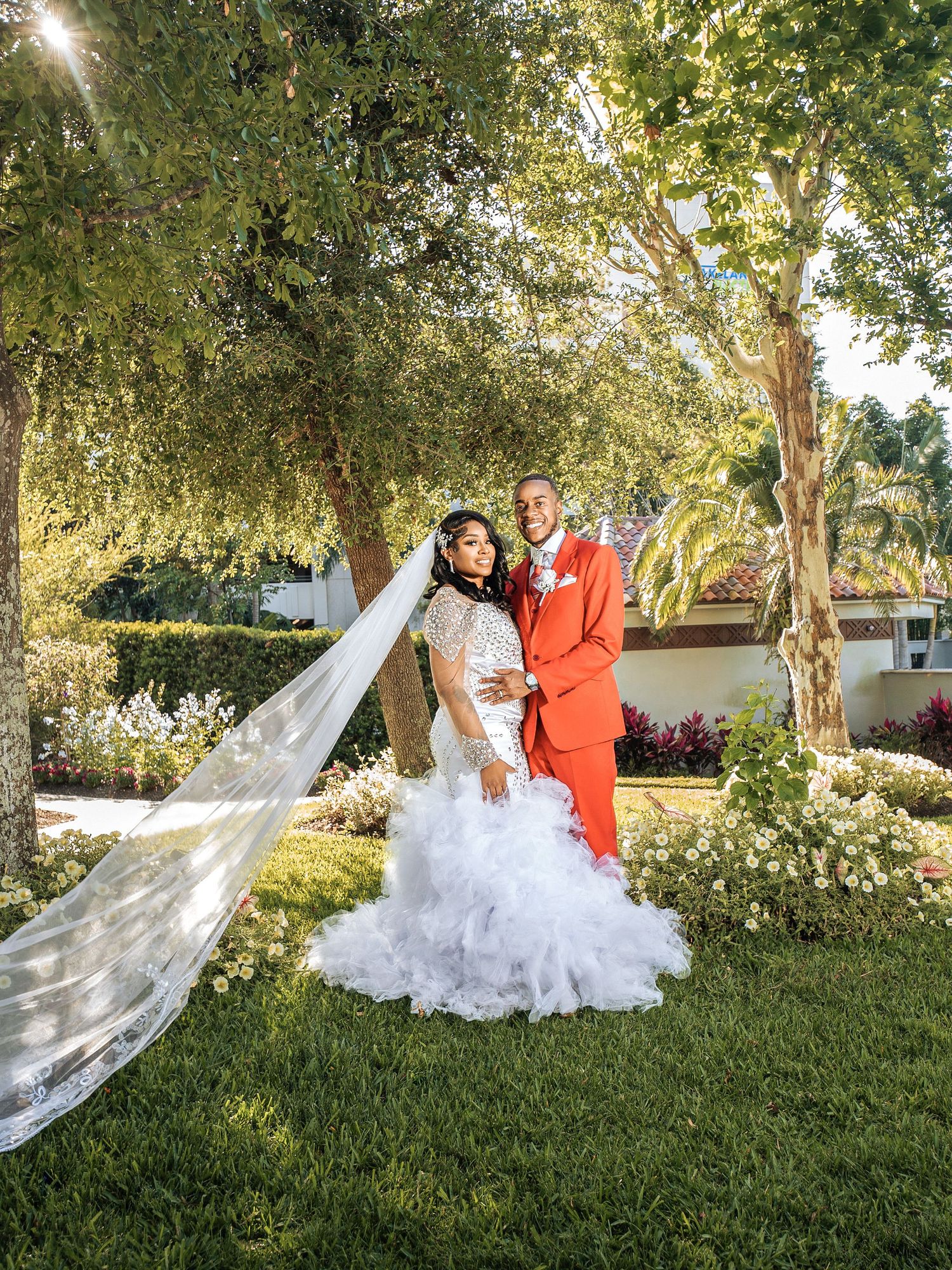 Couple in wedding attire embrace in a garden setting with lush greenery and sunlight filtering through the trees.