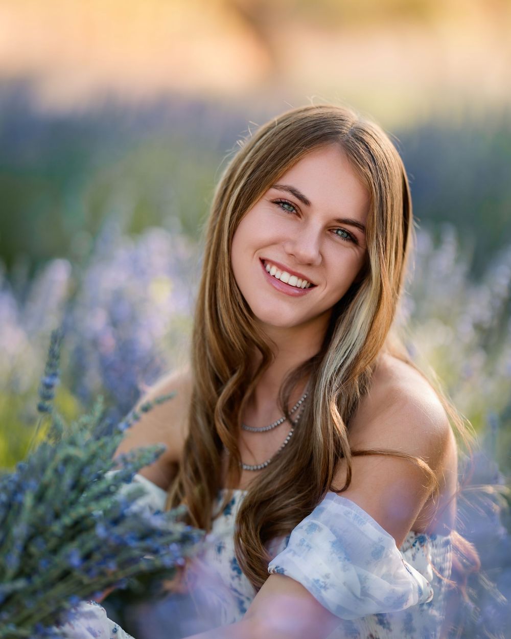 High school senior girl standing in a blooming lavender field at sunset, wearing a floral dress and smiling, photographed during golden hour