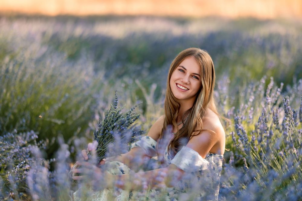 High school senior girl standing in a blooming lavender field at sunset, wearing a floral dress and smiling, photographed during golden hour