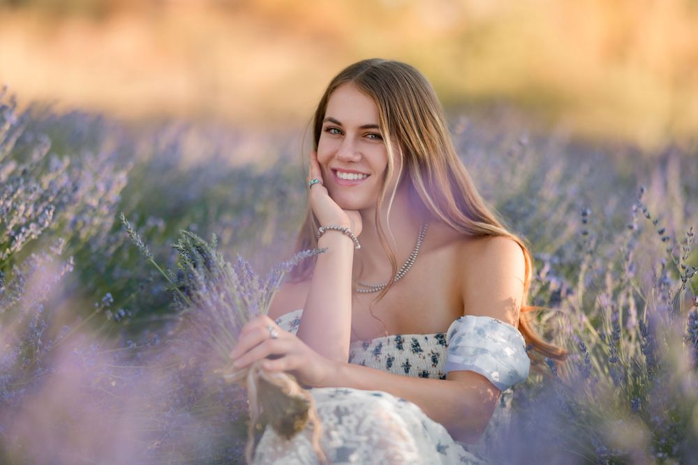 High school senior girl standing in a blooming lavender field at sunset, wearing a floral dress and smiling, photographed during golden hour