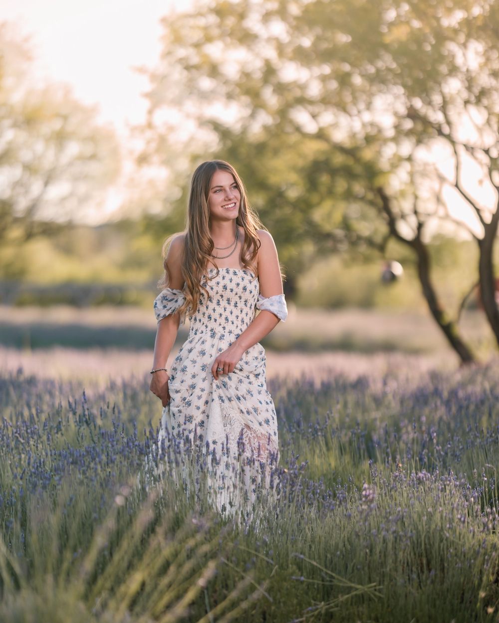 High school senior girl standing in a blooming lavender field at sunset, wearing a floral dress and smiling, photographed during golden hour