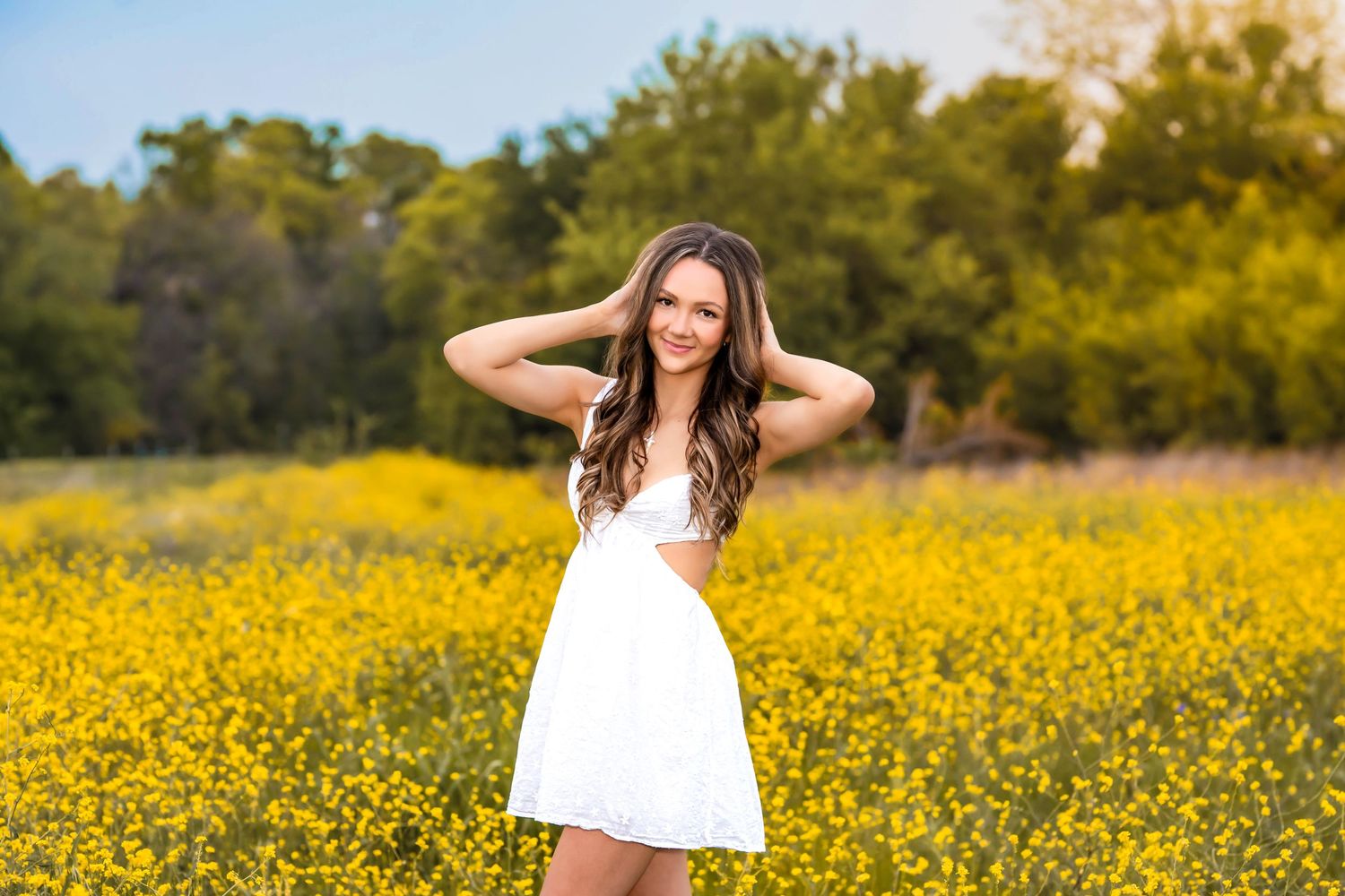 A woman wearing a white sundress poses in a vibrant yellow wildflower field on a sunny day.