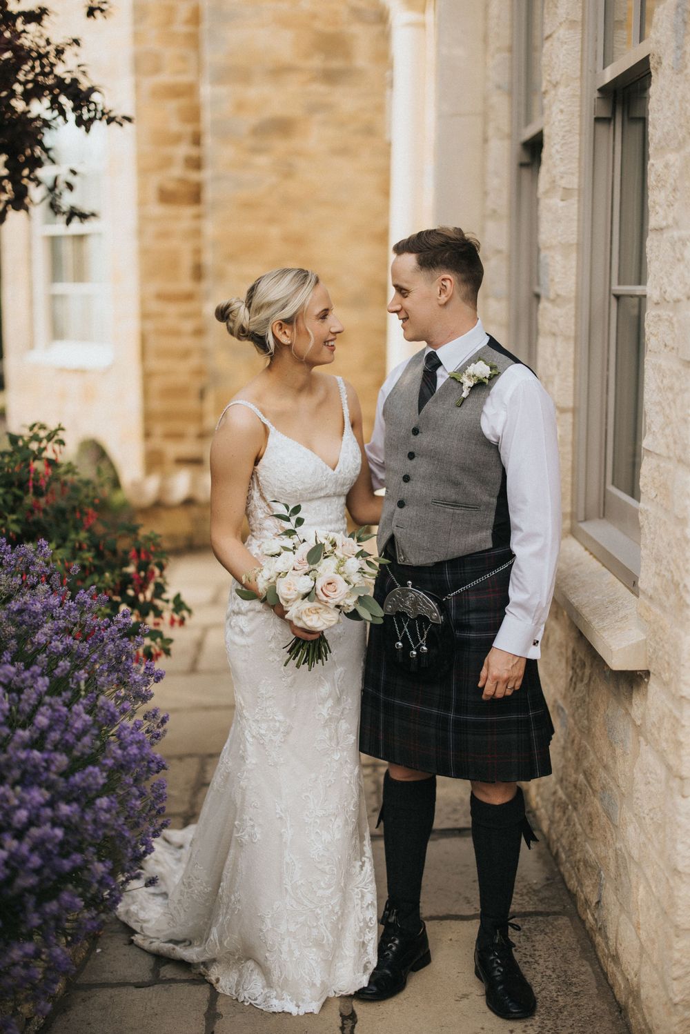 A bride in a lace gown and groom in traditional Scottish kilt attire share a moment against a stone building at Washingborough Hall.