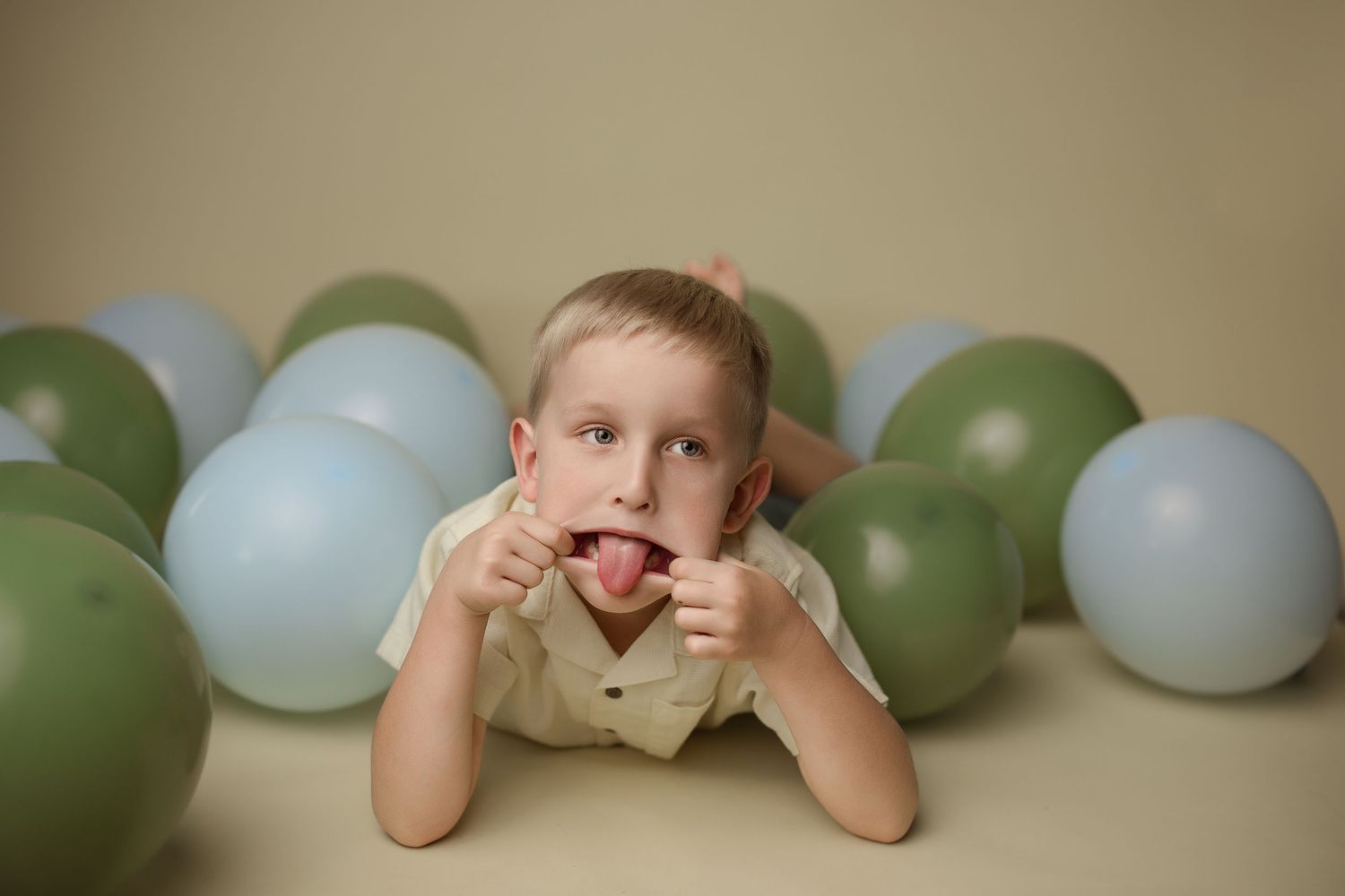 Child lying on floor making silly face surrounded by green and blue balloons against beige backdrop.