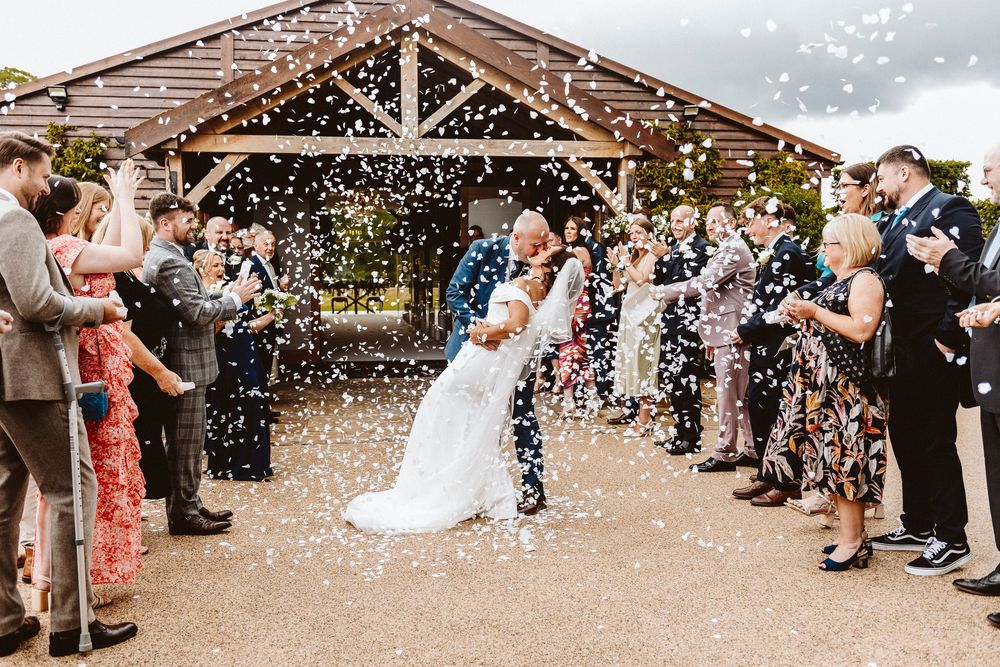 Wedding couple kissing under shower of confetti with guests lined up outside rustic venue.