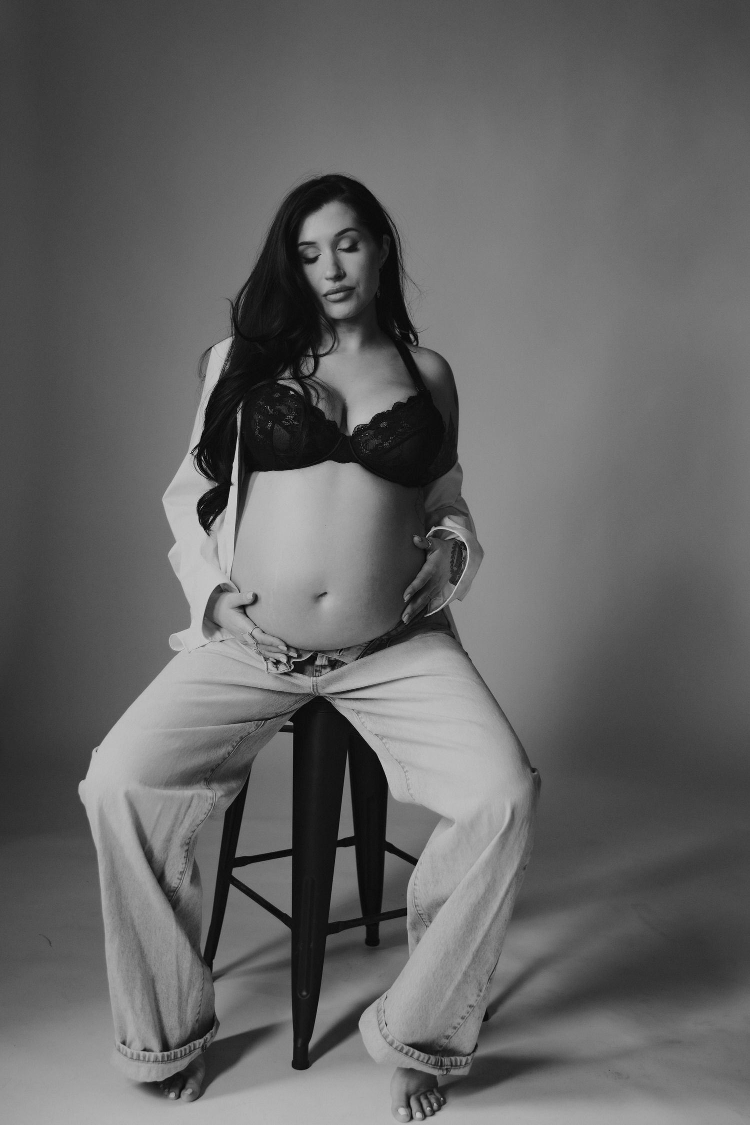Black and white artistic boudoir photo featuring a subject in black lingerie and loose pants seated on a stool.
