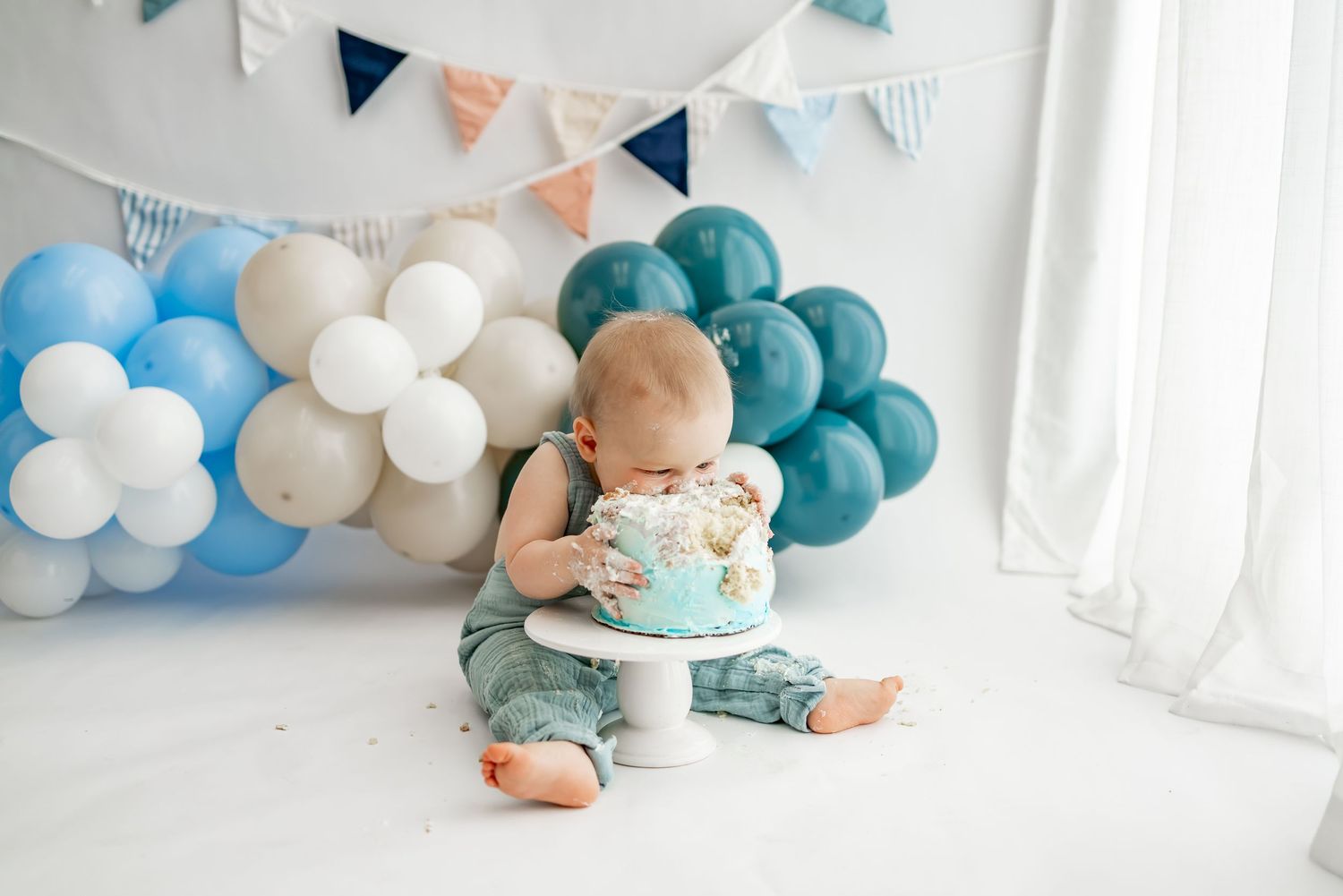 Baby in teal romper smashing cake during first birthday photoshoot with blue and white balloon decorations.