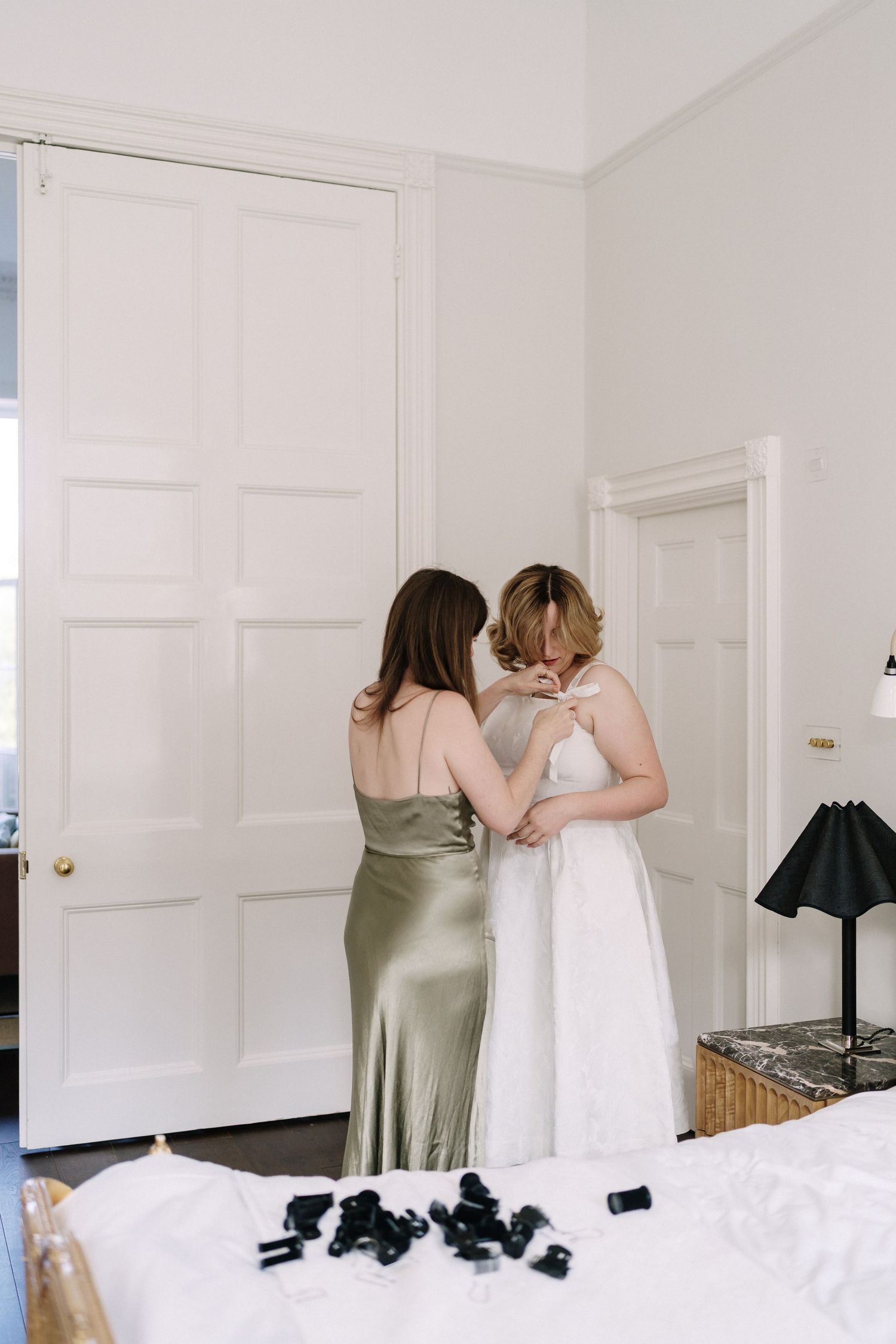Two people in formal attire help with dress preparations in a bright white bedroom interior.