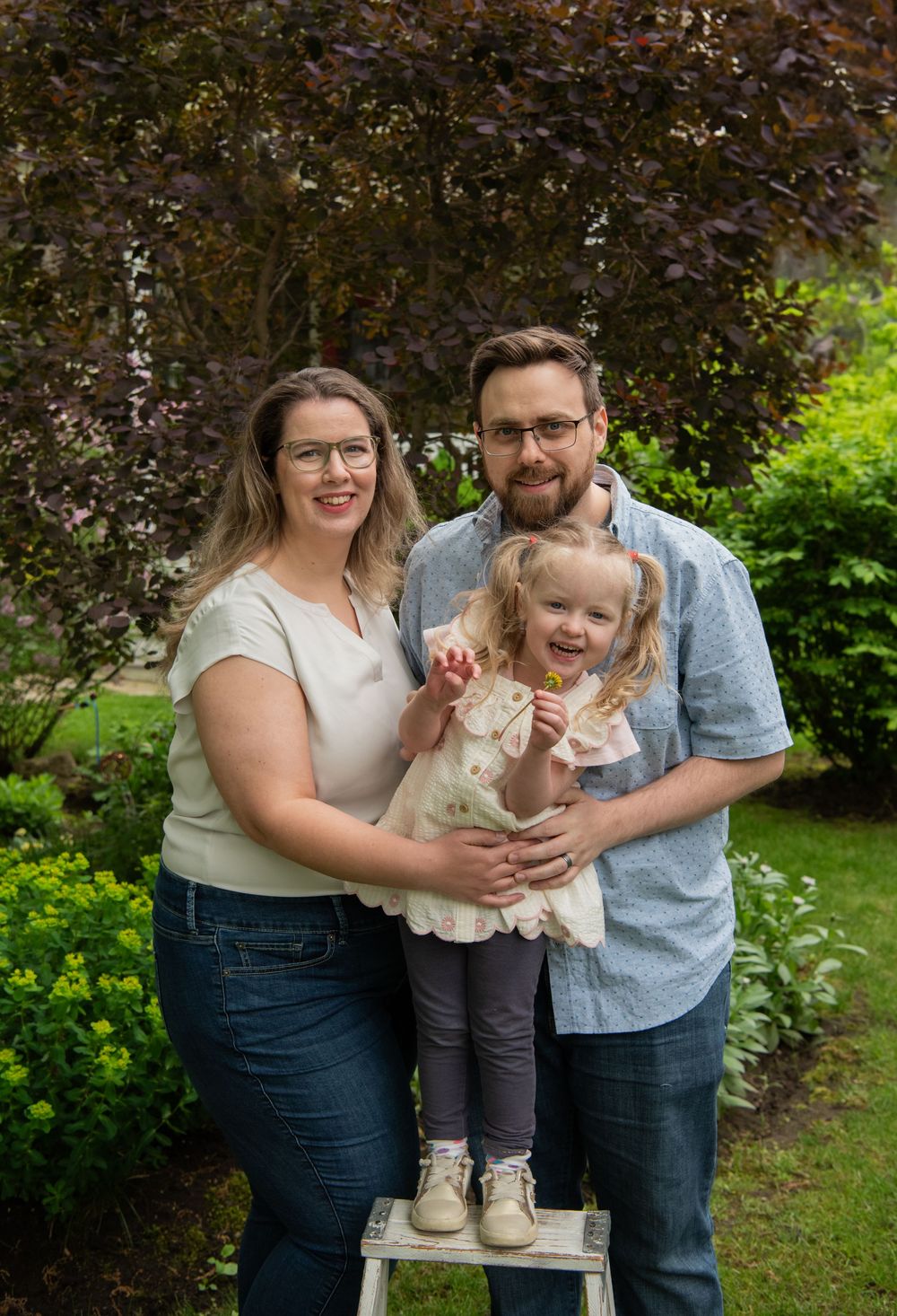 A happy family of three poses together in a garden setting with their toddler standing on a small stool.