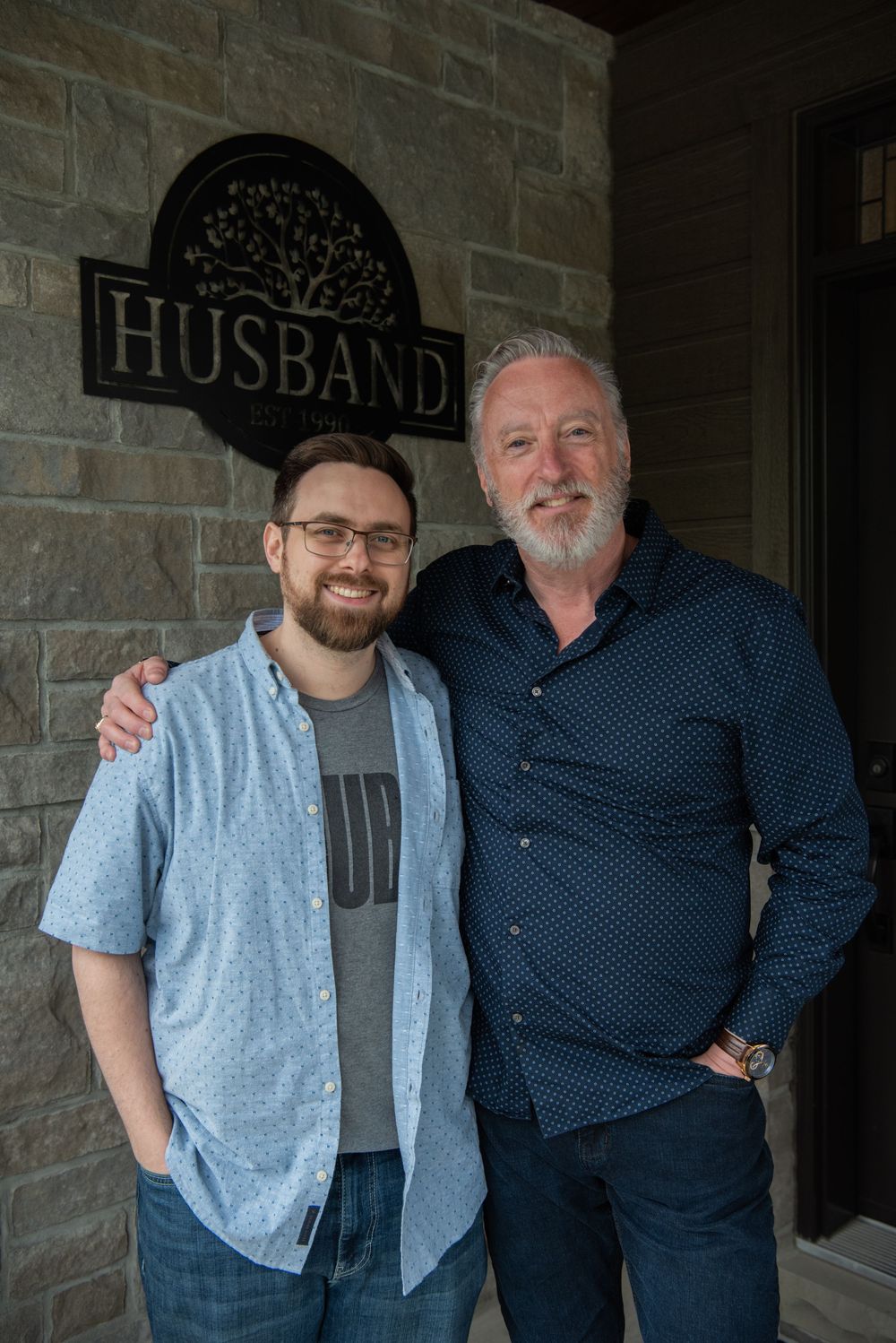 Two people stand together smiling outside a brick wall with a black decorative 'Husband' sign mounted above.