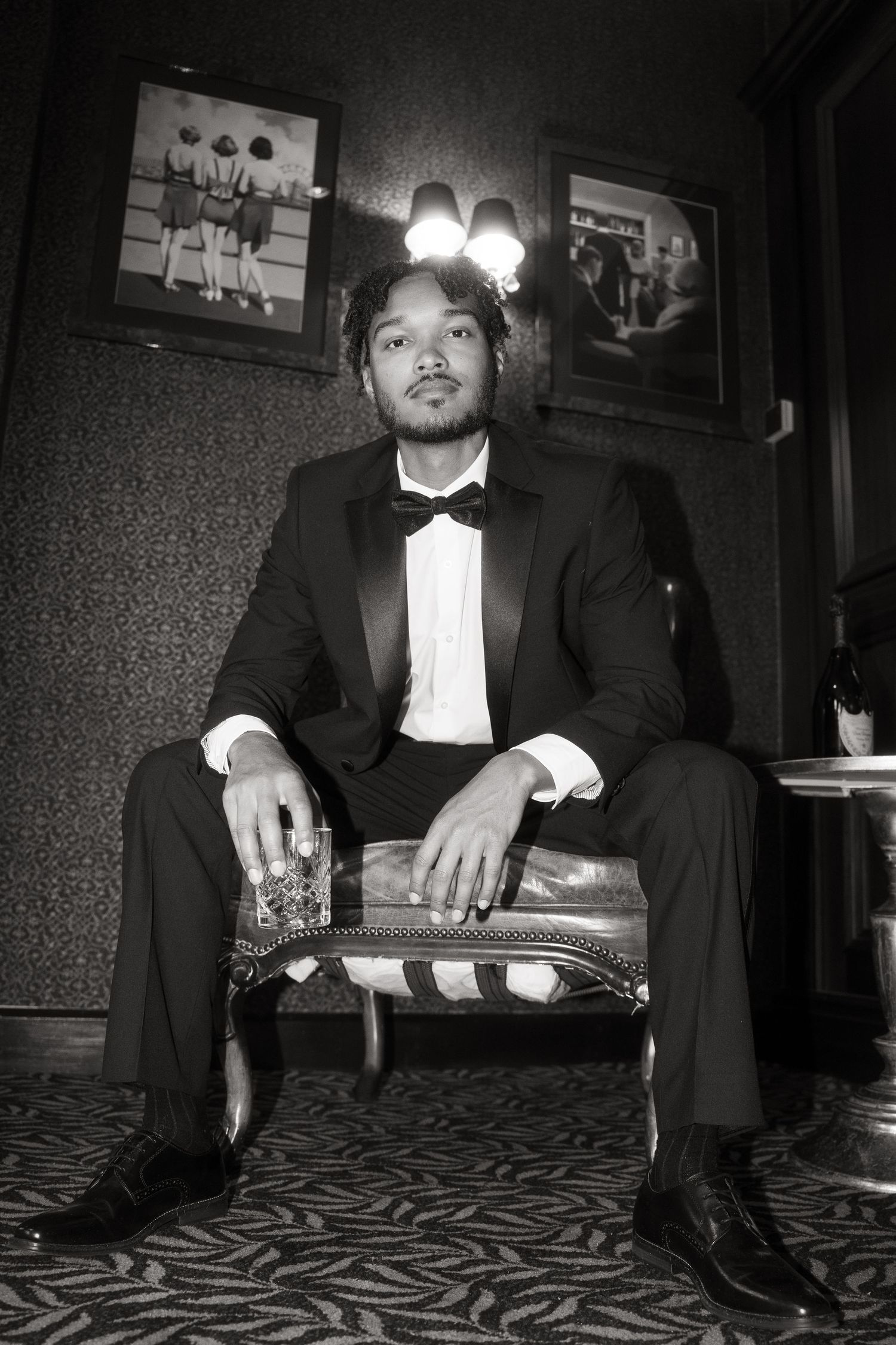 Black and white portrait of someone in a tuxedo sitting on a patterned chair in a dimly lit room.
