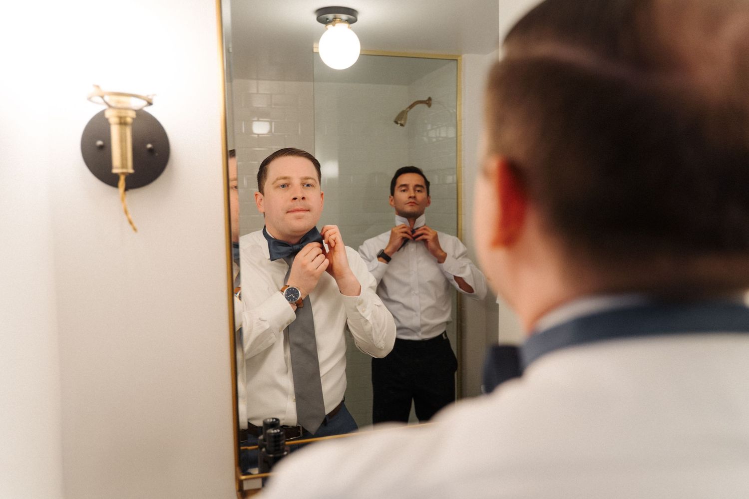 A person adjusts their necktie while looking in a bathroom mirror with warm lighting and minimalist decor.
