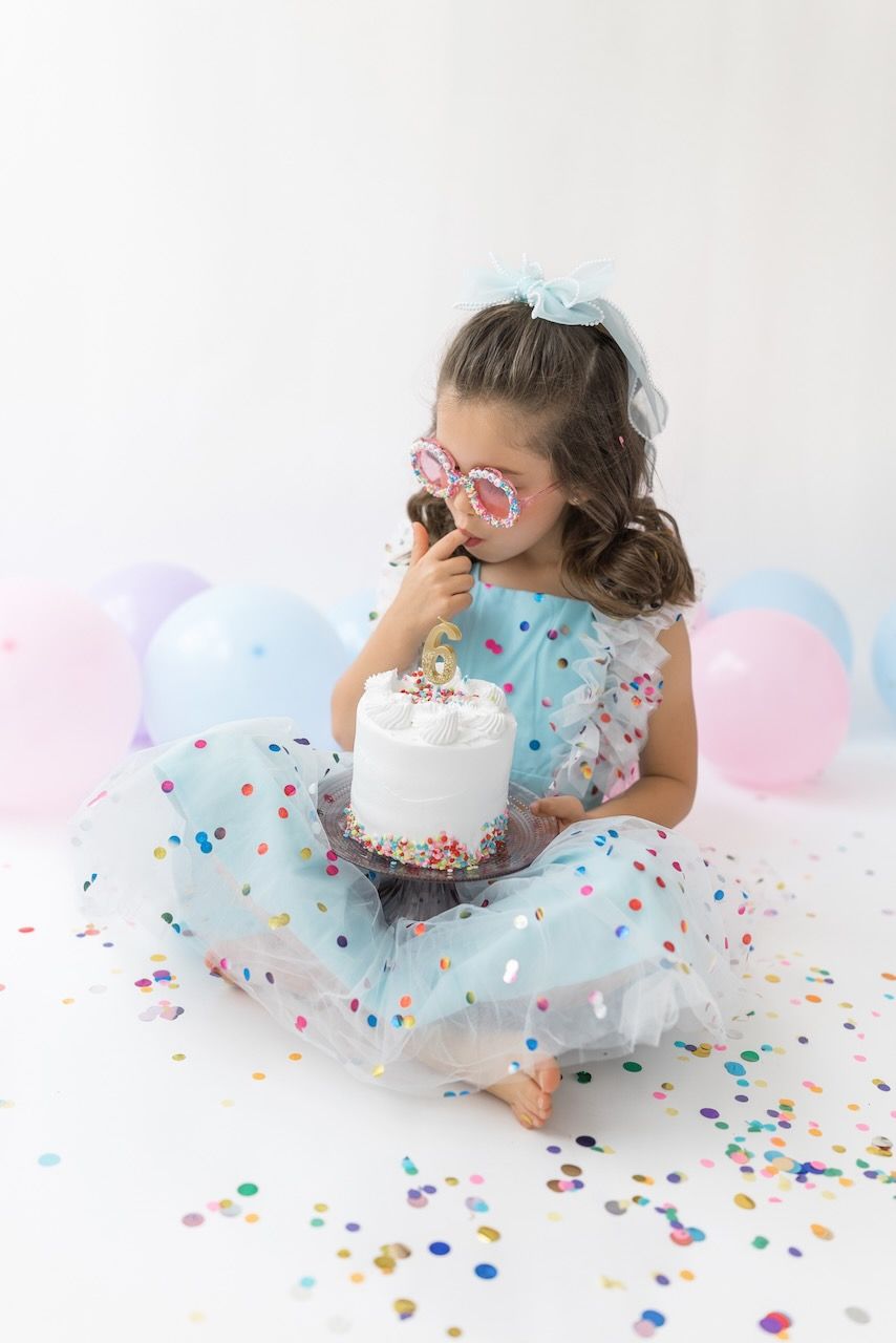 Girl wearing party glasses holding a mini birthday cake with pastel frosting and sprinkles.