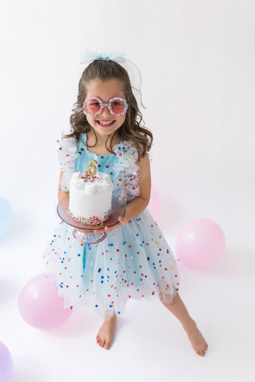 Girl wearing party glasses holding a mini birthday cake with pastel frosting and sprinkles.