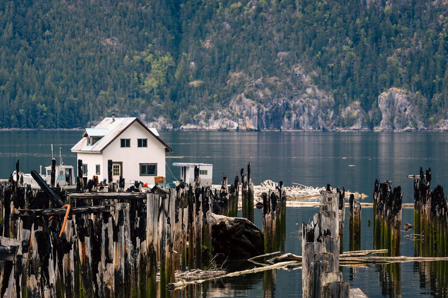 White waterfront house stands among weathered wooden pilings on the Pacific Ocean in Bella Coola with mountains in the background.