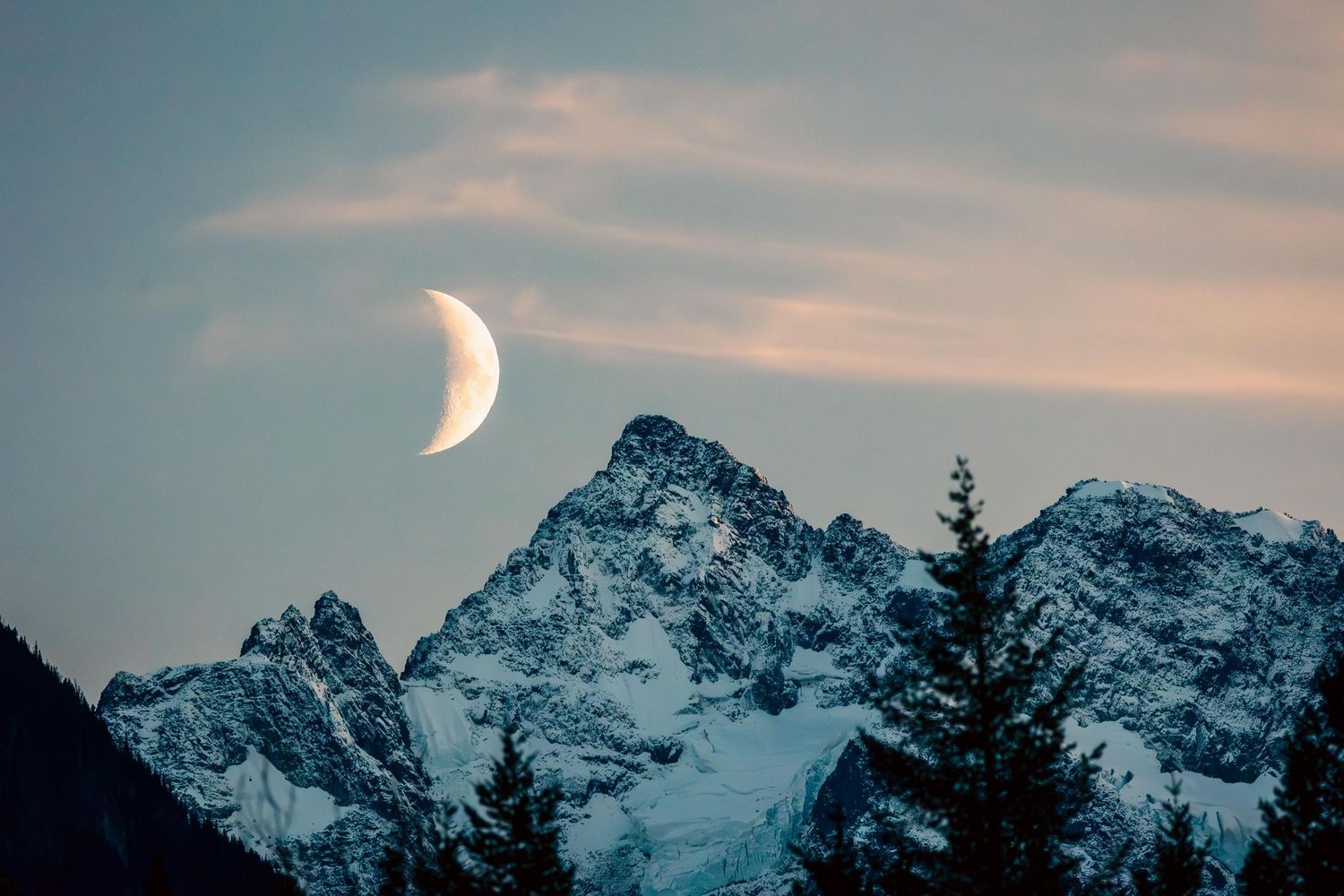 A crescent moon rises over snow-capped mountain peaks with silhouetted evergreen trees in the foreground at dusk in the Bella Coola Valley.