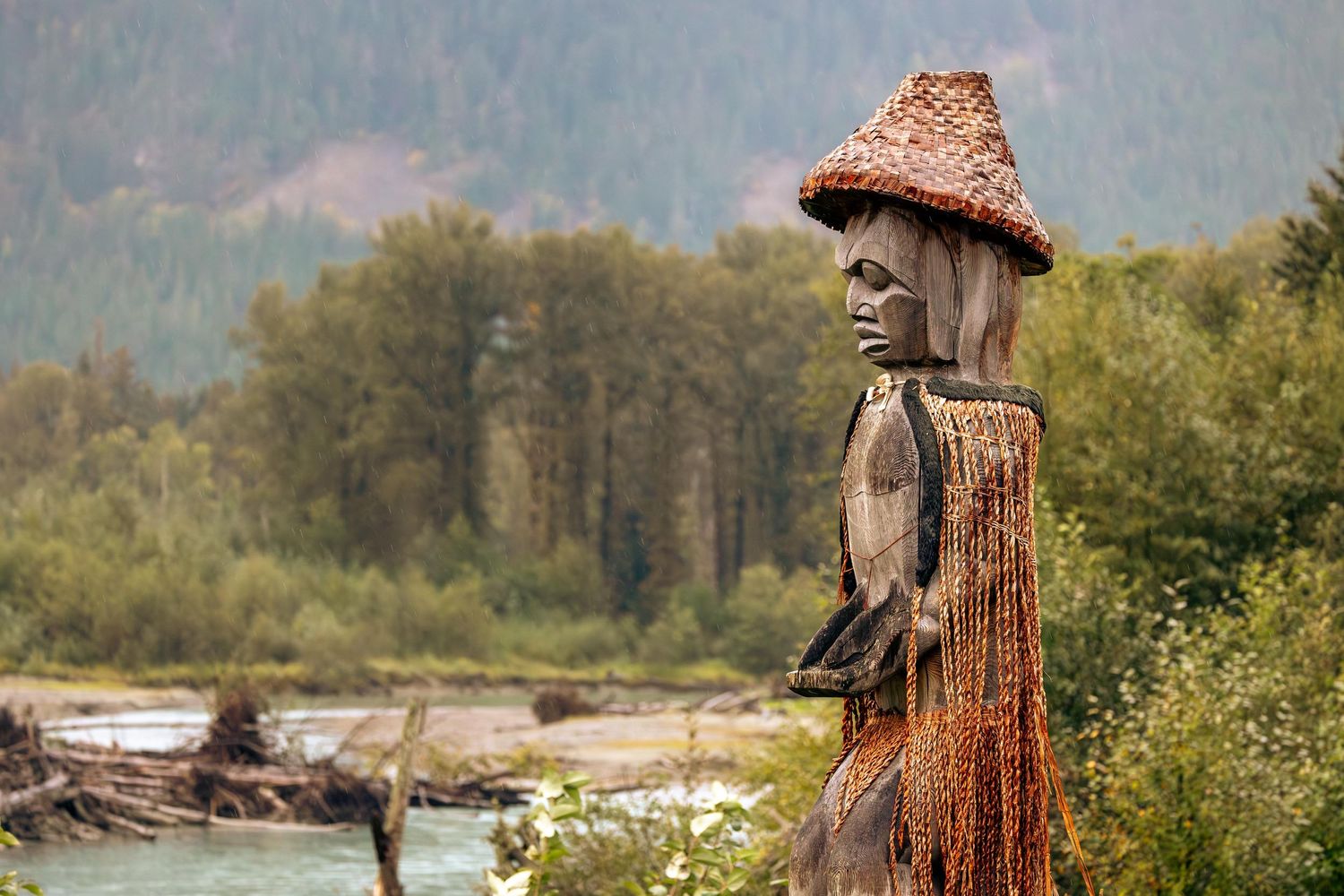 Nuxalk totem pole carving wearing a cedar hat stands near a riverbank in the Bella Coola Valley.