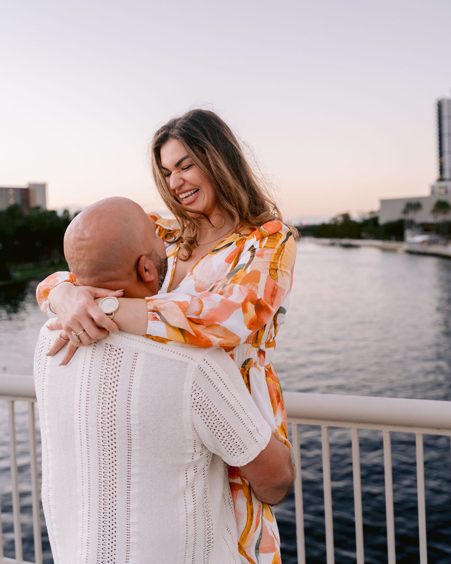 A couple shares a joyful embrace while standing on a waterfront pier at sunset.