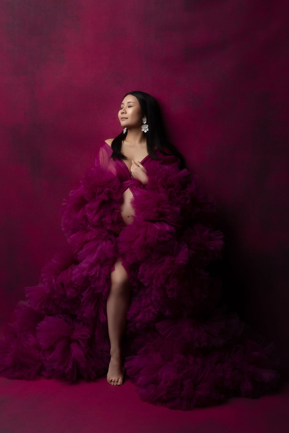 Figure in a flowing burgundy tulle gown poses dramatically against a dark red studio backdrop.