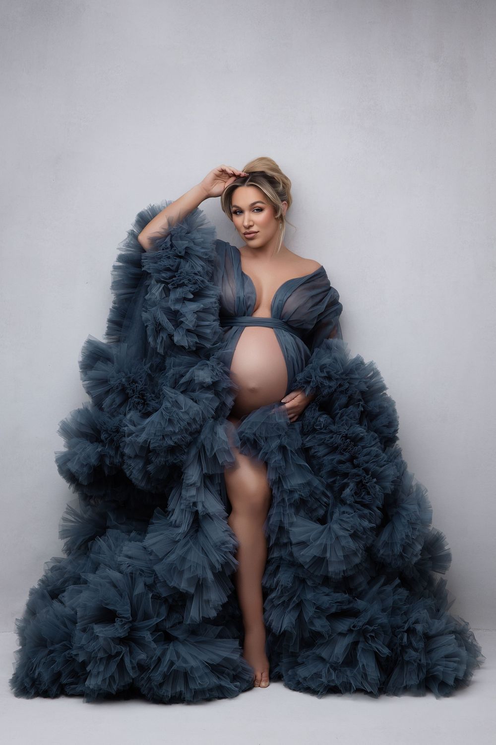 A maternity photo featuring an elegant teal tulle gown with dramatic ruffles against a light gray background.