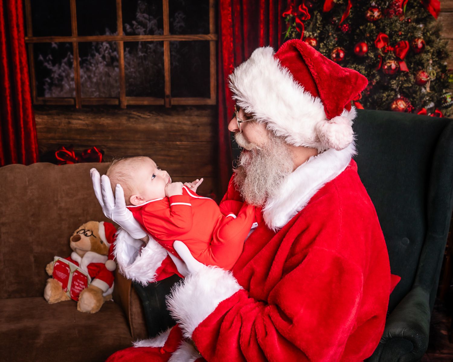 Santa Claus holds a small baby in red clothing against a warm Christmas backdrop with decorations.