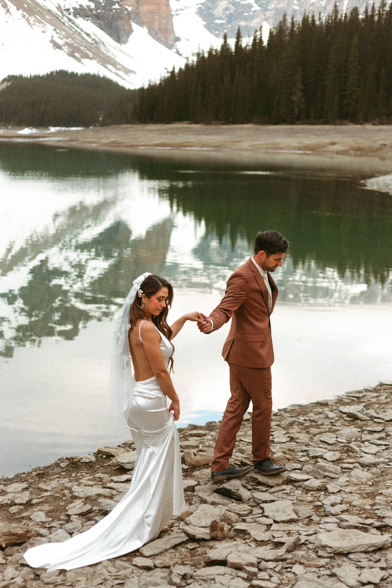 Wedding couple holding hands on rocky shoreline beside mountain lake with snow-capped peaks and evergreen forest reflections.