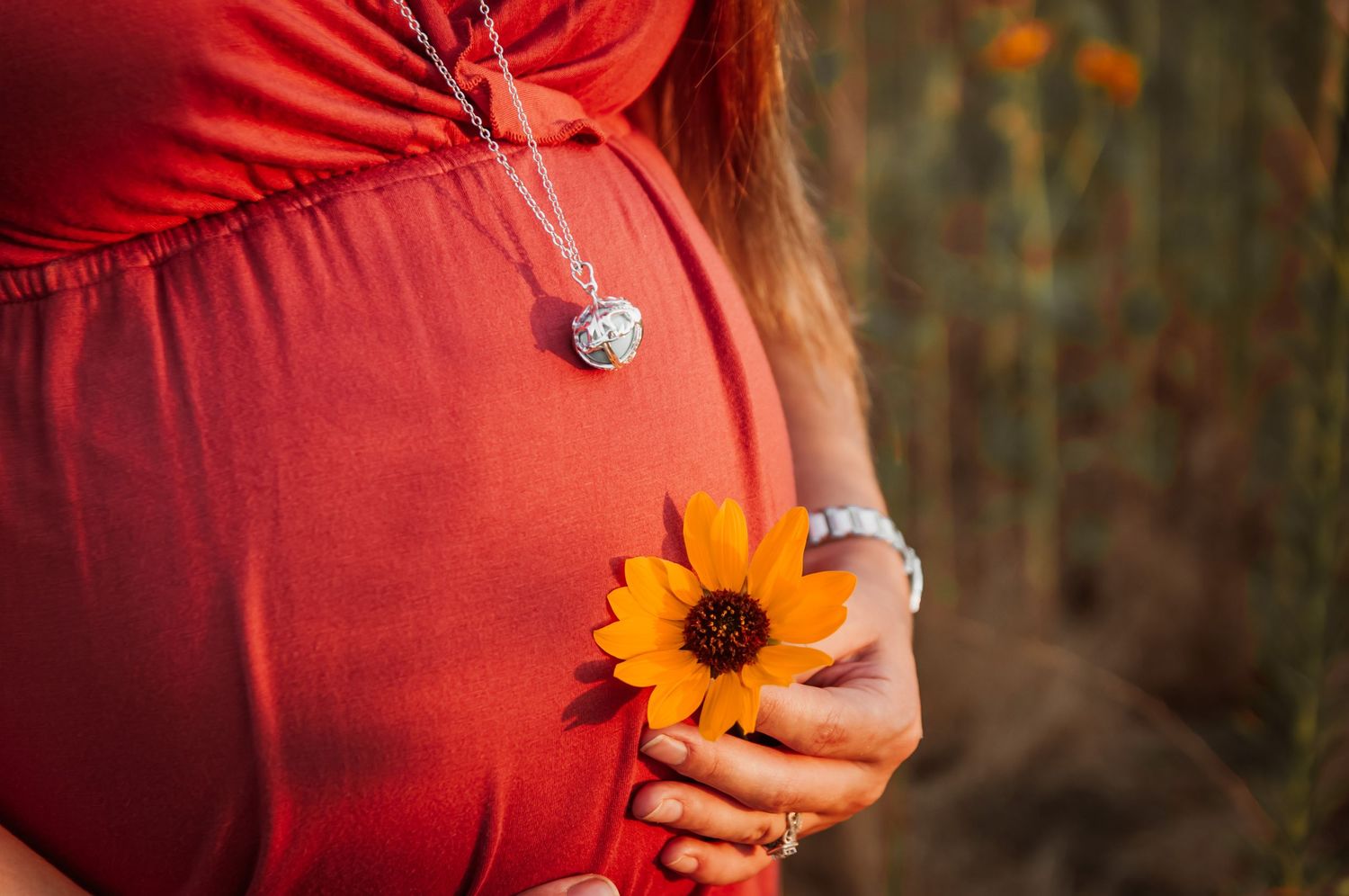 A hand holds a bright orange sunflower against a red maternity dress while wearing a heart-shaped necklace.
