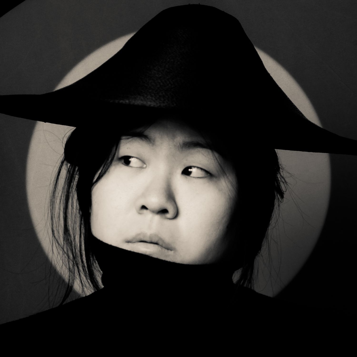 Artistic black and white portrait with dramatic lighting features a person wearing a wide-brimmed black hat against light backdrop.