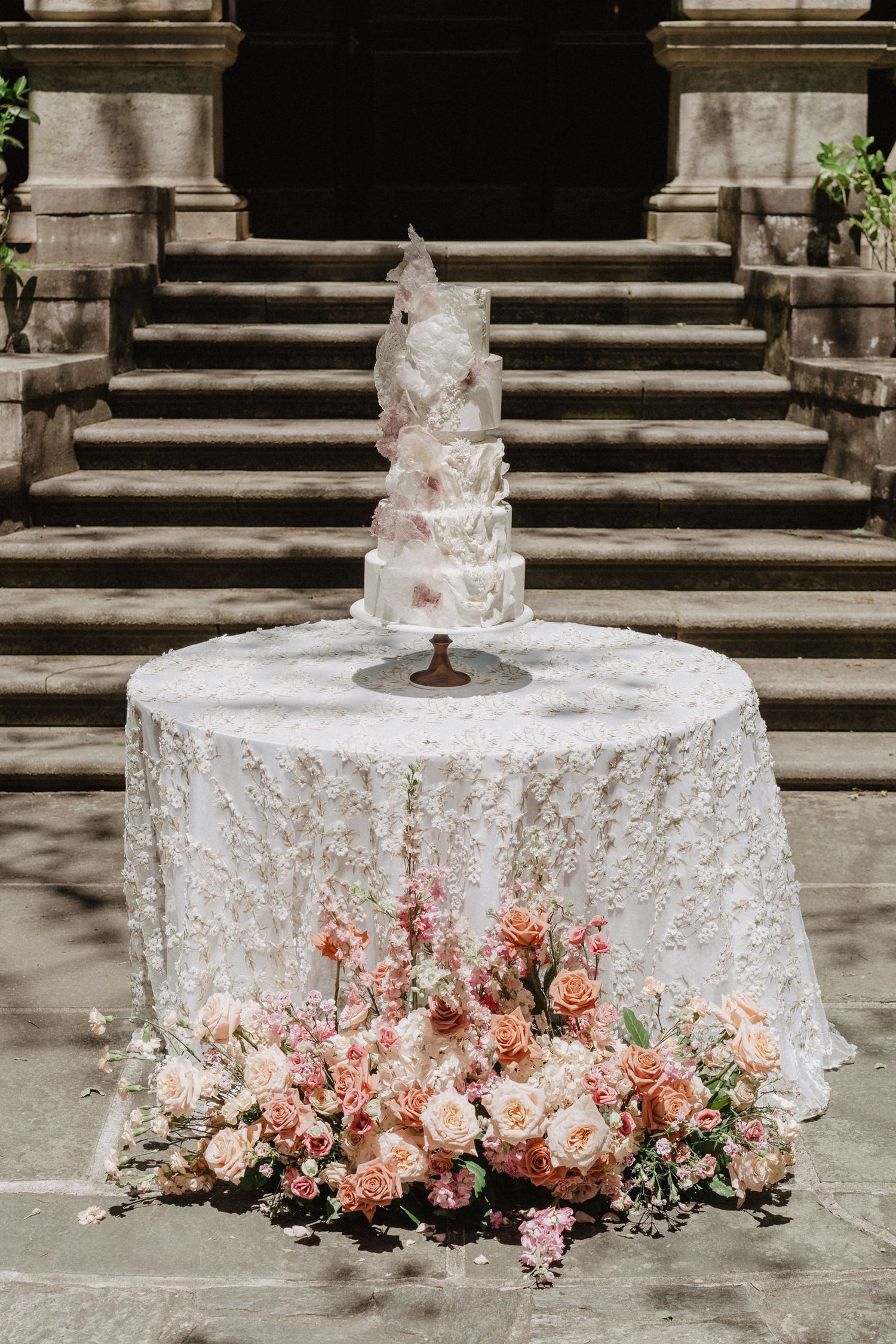 Elegant white wedding cake with coral floral arrangement on sequined tablecloth against stone steps.