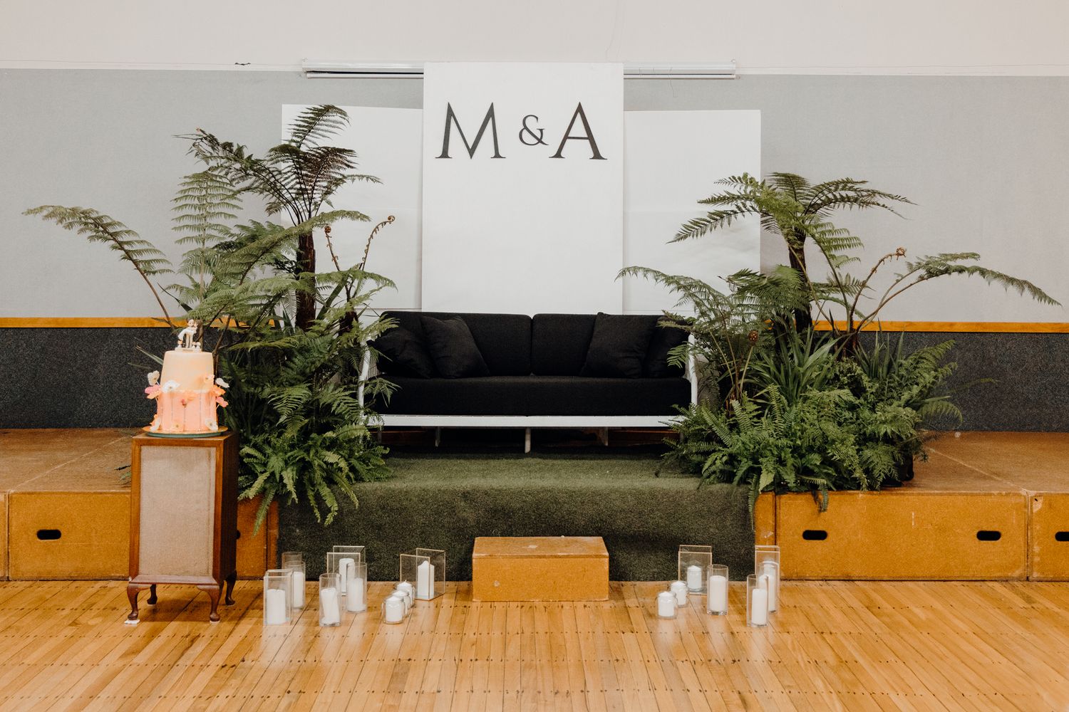 A wedding reception setup with M&A sign, black couch, and potted ferns in a wood-floored room.