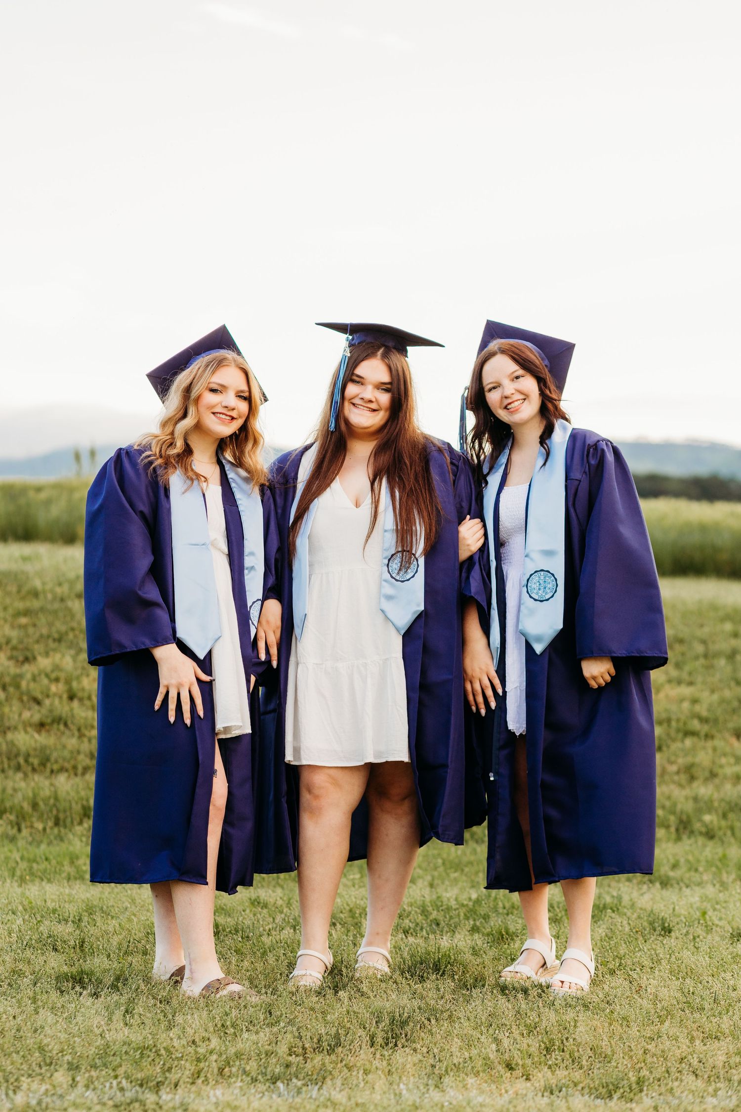 Three graduates in navy blue caps and gowns stand together smiling on grassy field with mountains in background.