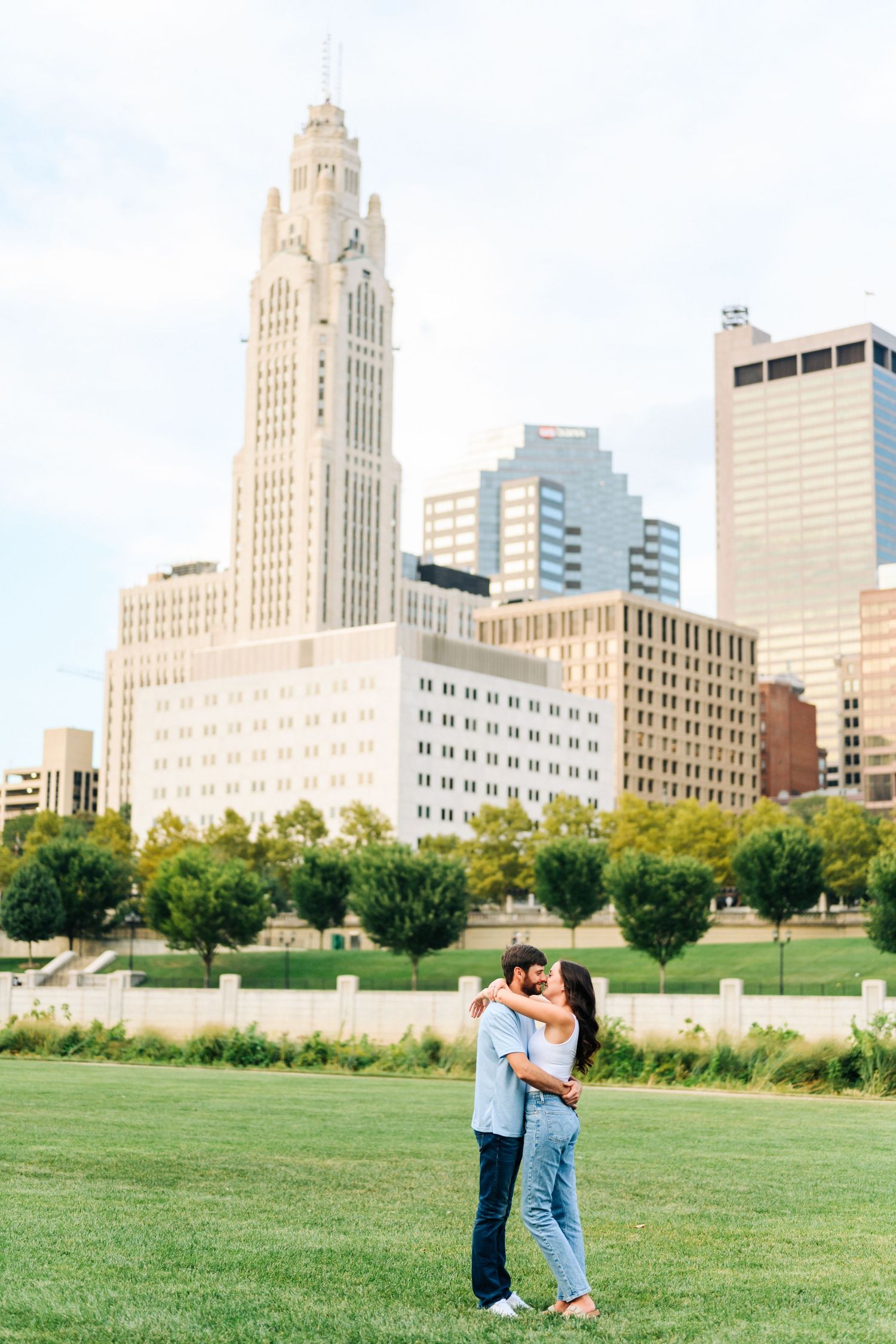 A couple embraces on a green lawn with Columbus, Ohio's LeVeque Tower and downtown skyline visible in the background.