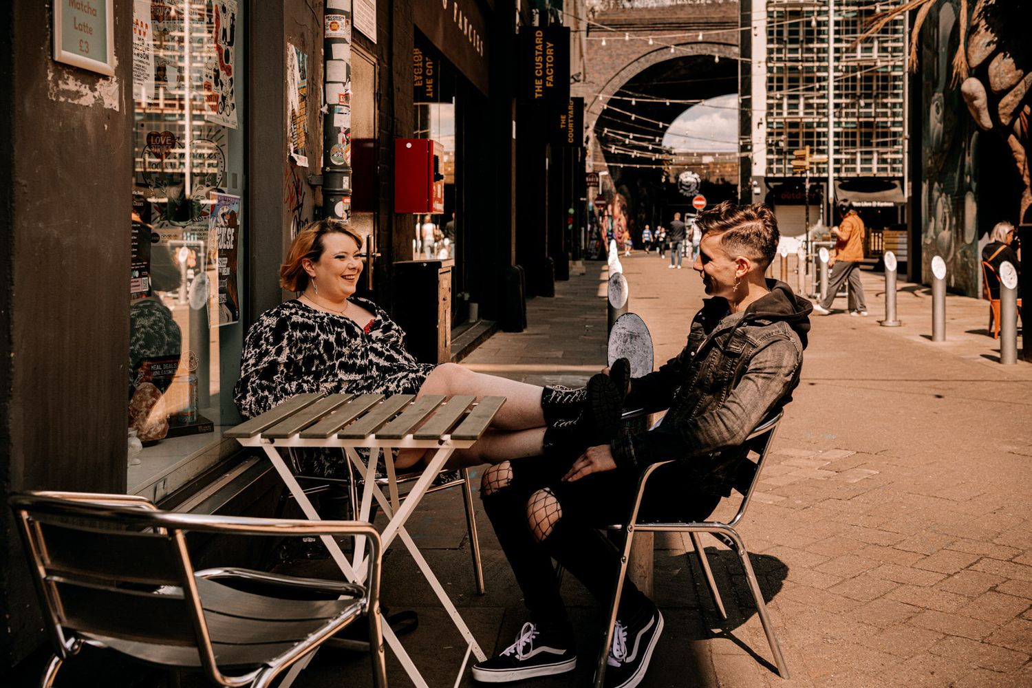 Two people enjoying coffee at an outdoor cafe table along a busy city street with arched architecture.