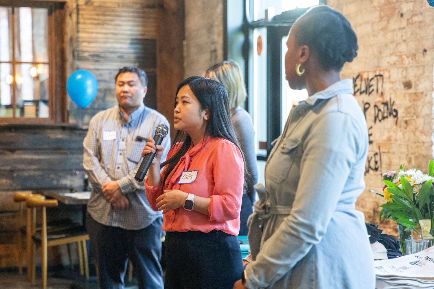 Three coworkers engage in animated conversation during a casual meeting in an industrial-style office space with exposed brick walls.