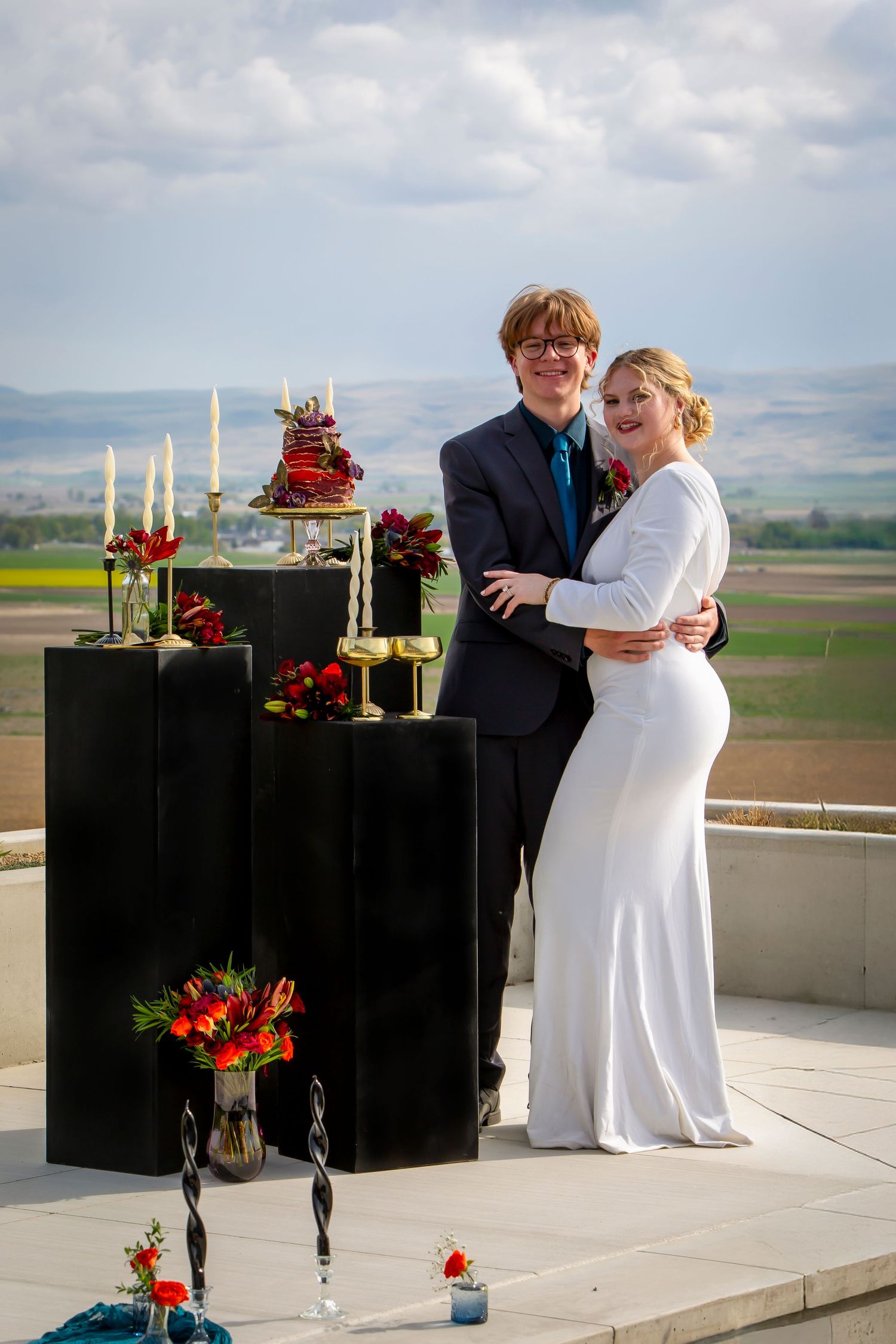 wedding couple posing by black pillars with red, orange and purple florals, white candles, gold accents, and a red and purple wedding cake