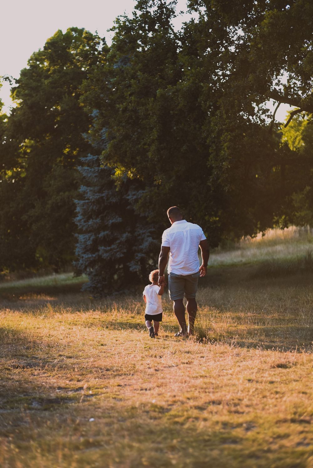 A parent and child walk together on a dry grass field at sunset, with green trees and a blue spruce in the background.