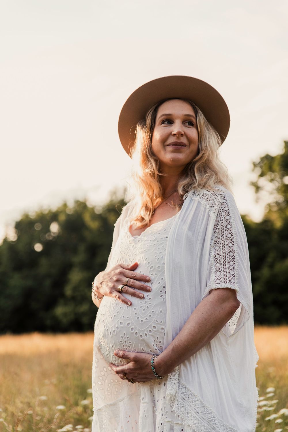 Expectant mother stands in golden field at sunset wearing white lace dress and brown hat while cradling baby bump.