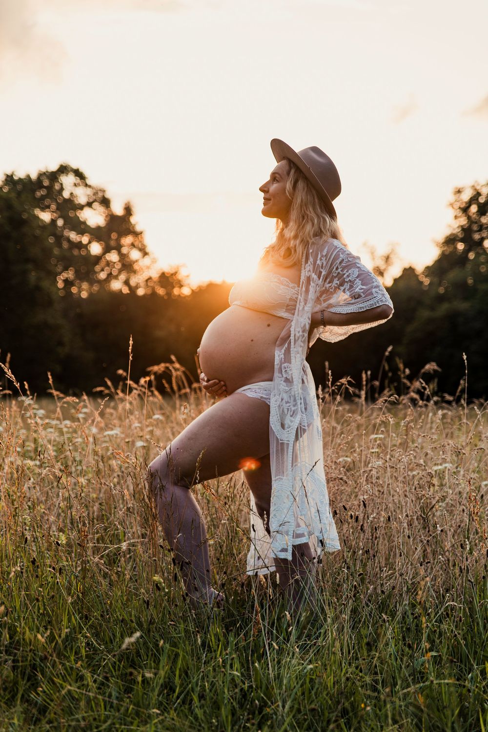 Silhouette of pregnant woman in hat and lace robe standing in golden meadow at sunset.