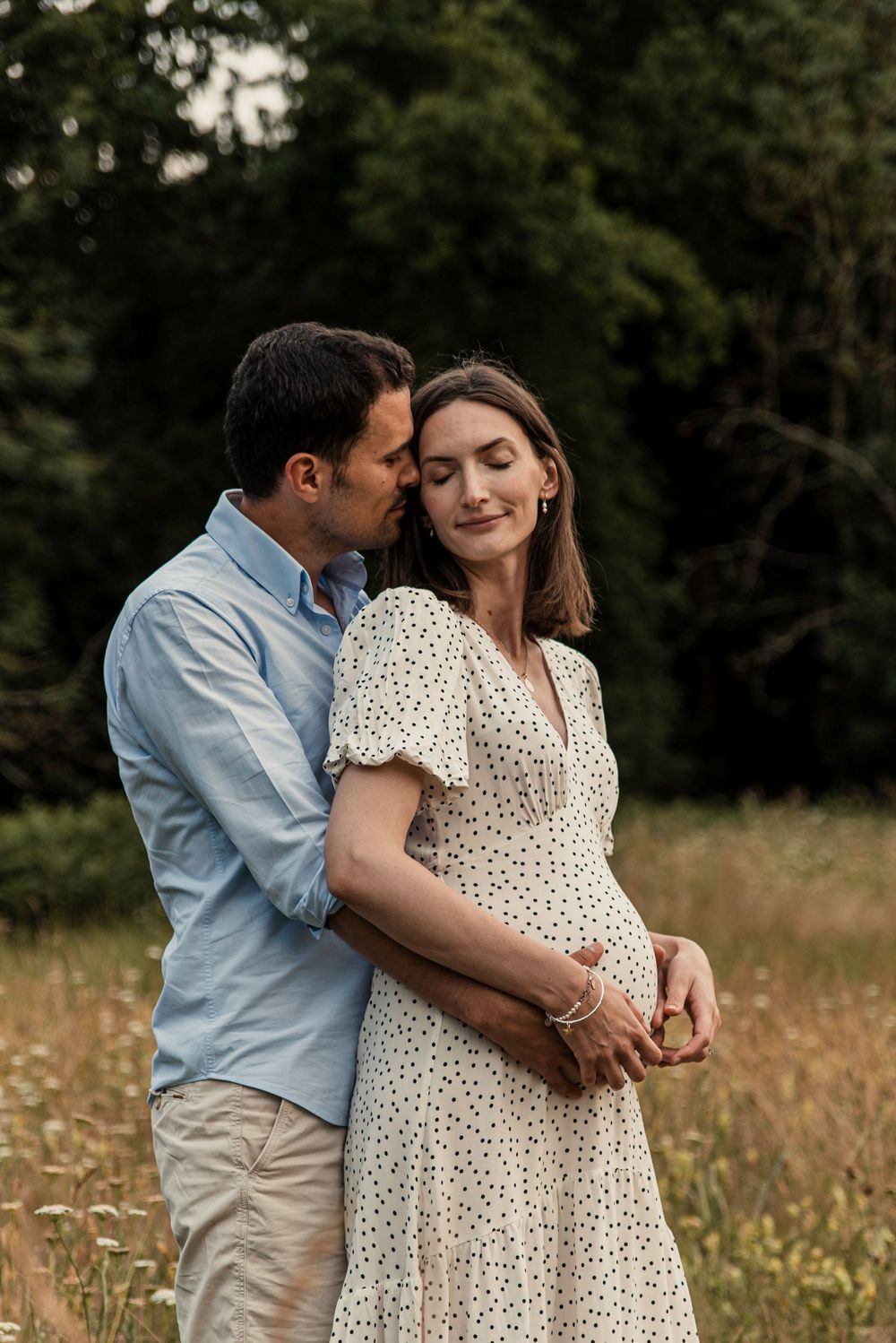 Couple shares intimate moment in field of wildflowers as sun sets behind trees, displaying tender affection and joy.