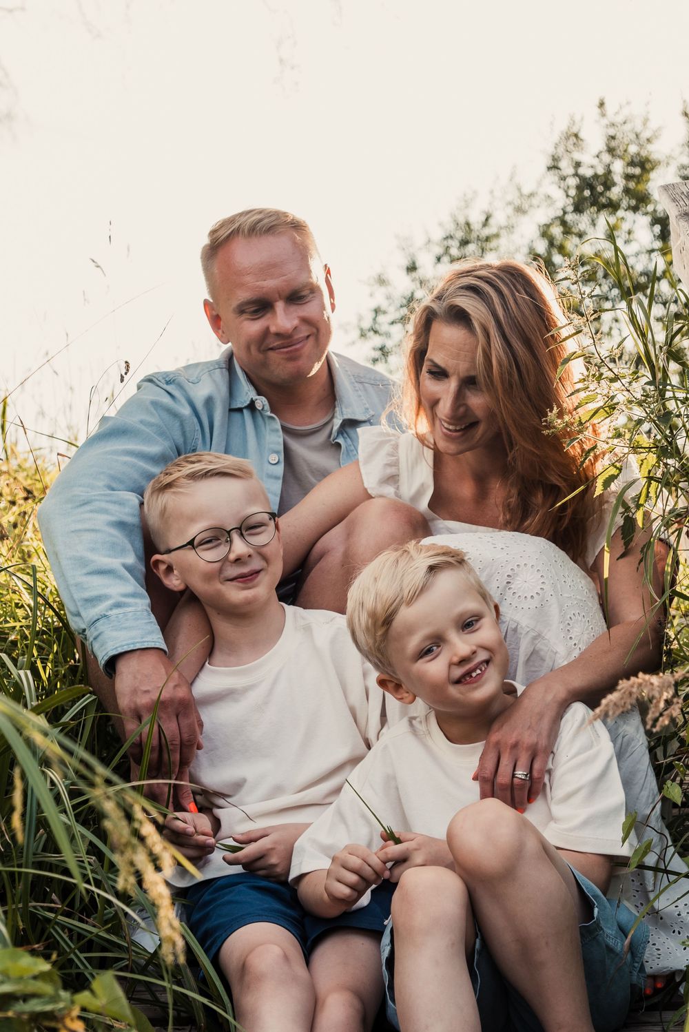 A loving family sits together outdoors in tall grass during golden hour, all wearing casual white and denim outfits.
