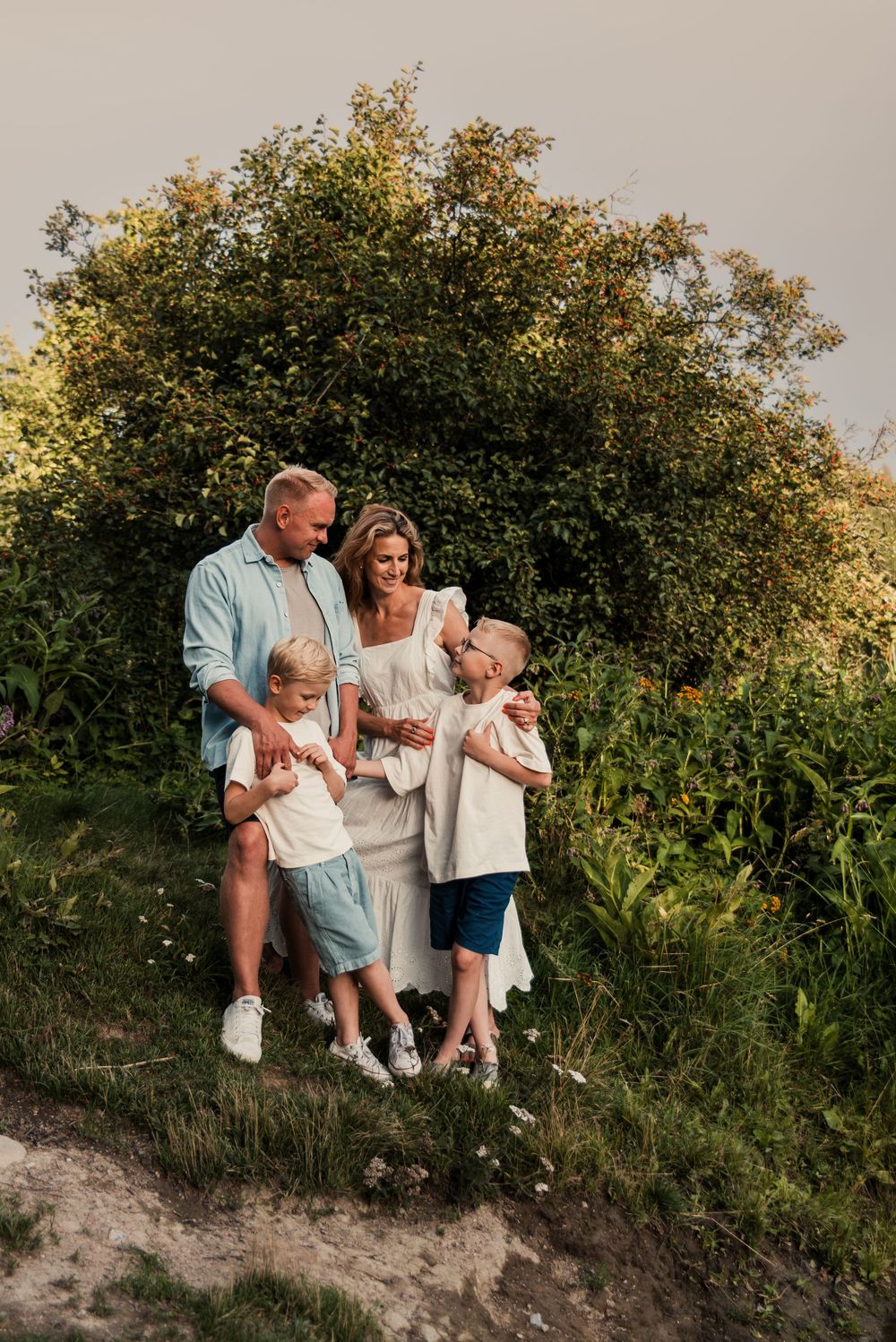 A family stands together outdoors on a sandy path against a lush green tree during golden hour sunlight.